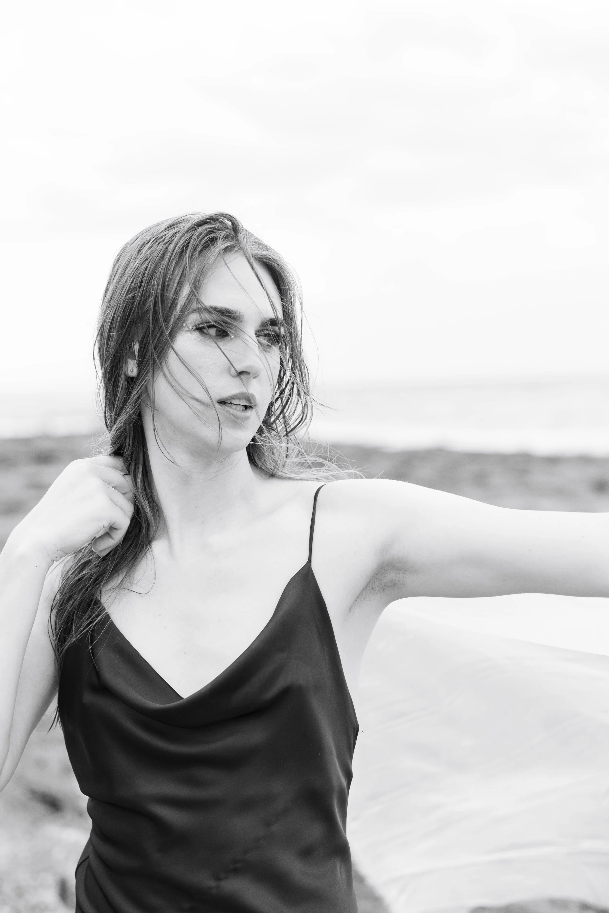 A woman in a cowl nexk dress standing outdoors by the water, with her arm extended outward and hair blowing in the wind, captured in black and white.