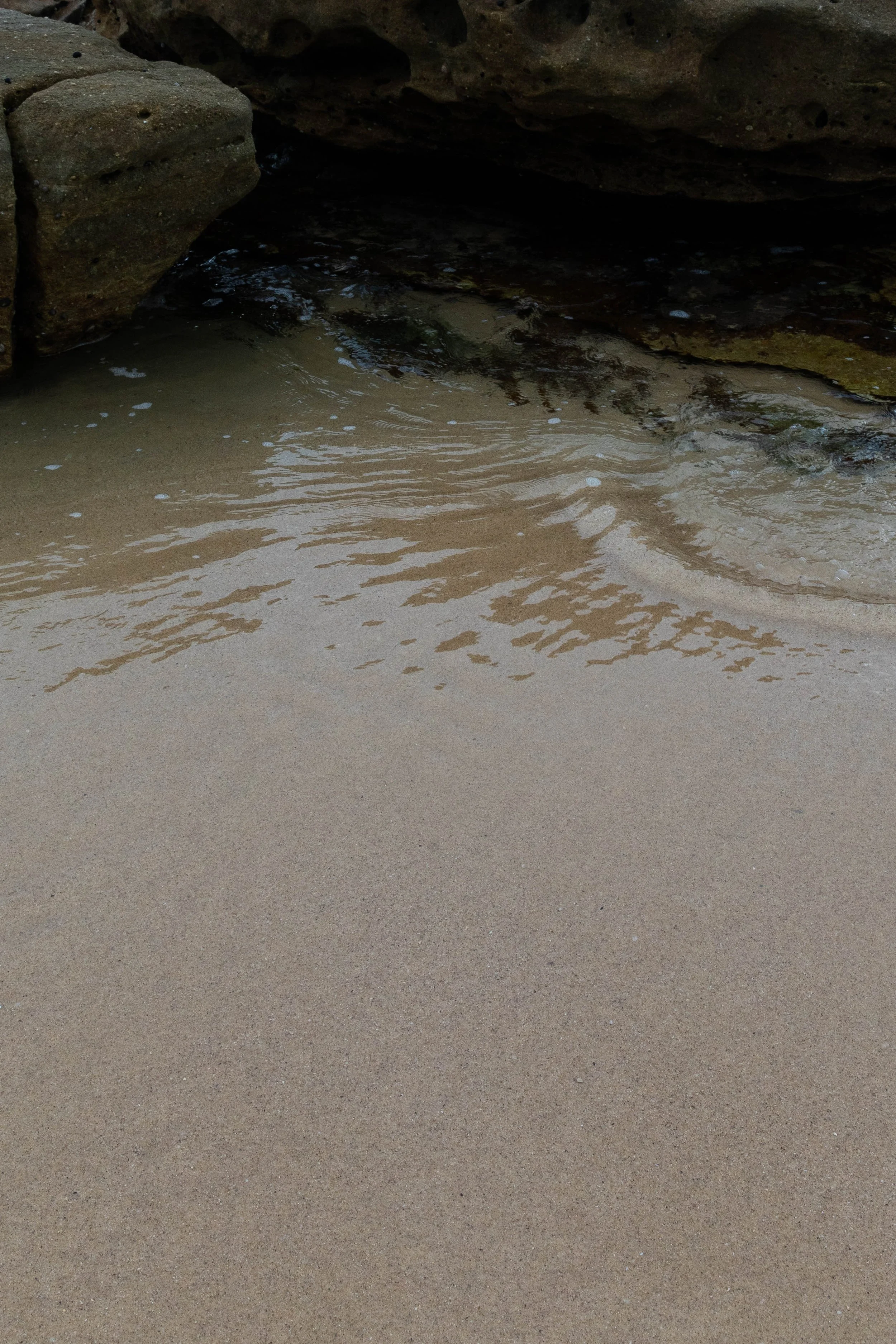 Sand and water at the base of rocks, displaying gentle waves and foam.