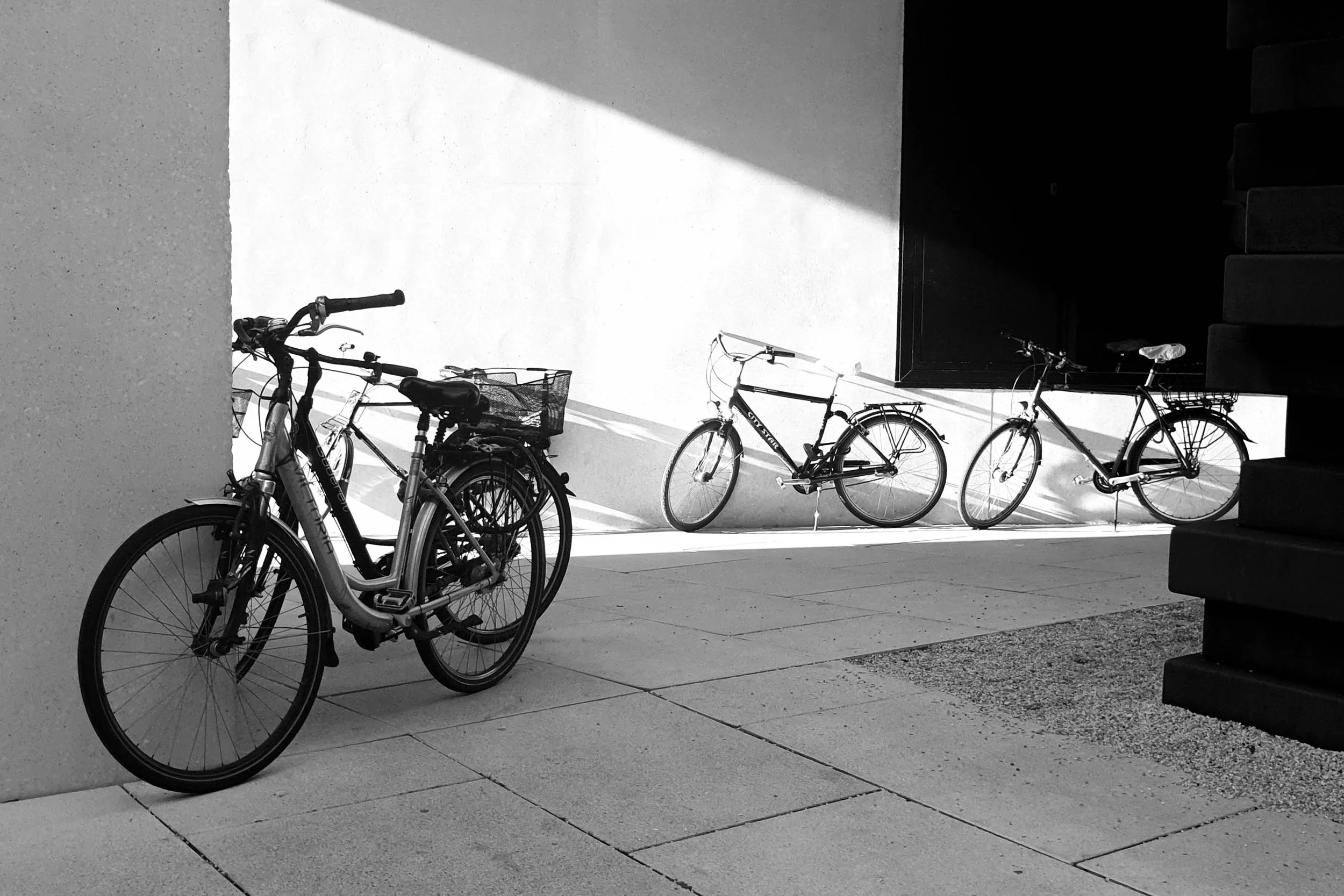 Three bicycles parked against a white wall, with shadow cast by sunlight, and concrete paving in the foreground, black staircase on the right side.