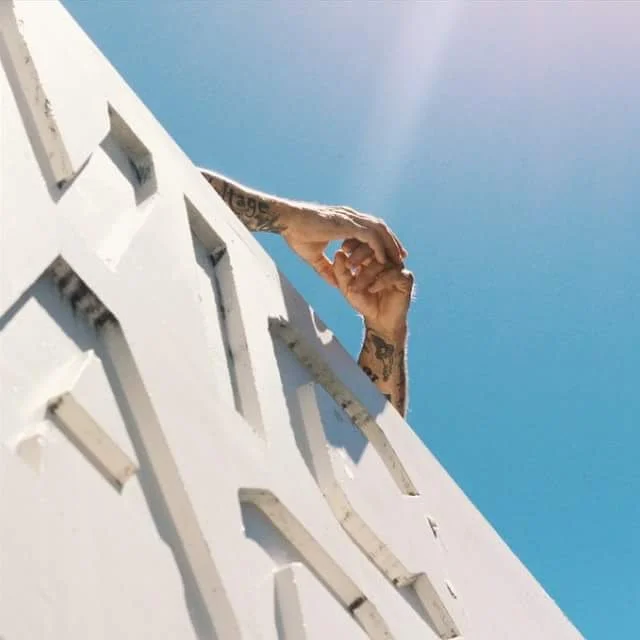 A person with tattoos extending their arm over a white railing against a clear blue sky.