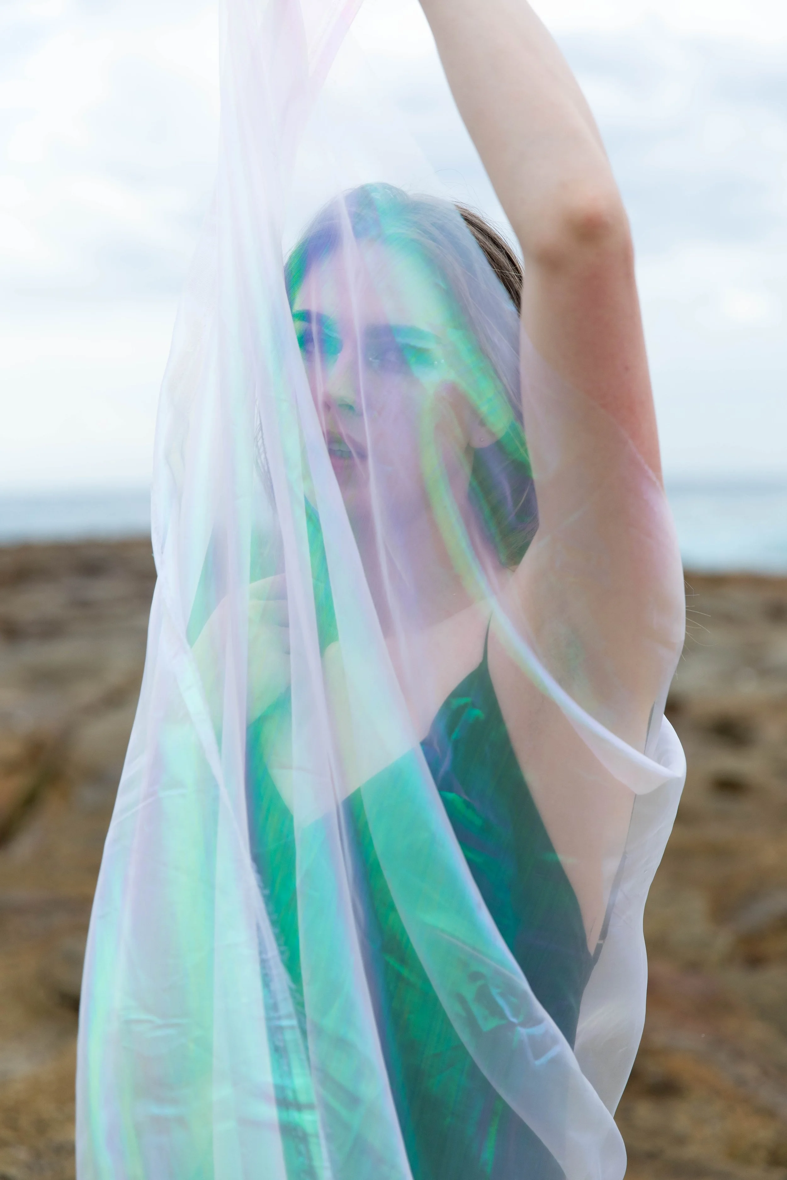 A woman stands outdoors near a beach, holding a sheer, iridescent fabric above her head while smiling.