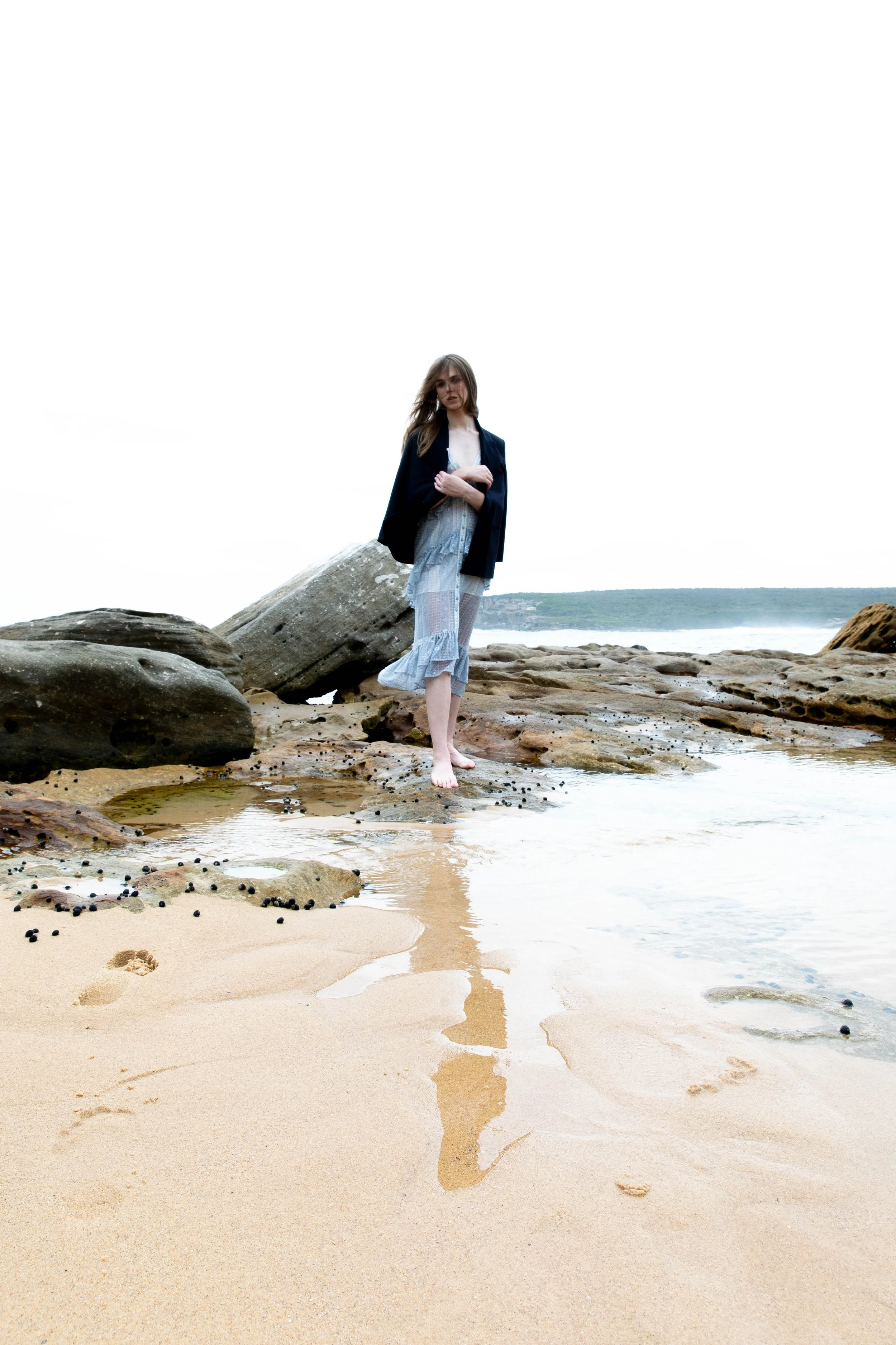 A woman standing barefoot on a sandy beach with rocks, wearing a black jacket over a white top and a light blue skirt, looking at the camera with an overcast sky and water in the background.