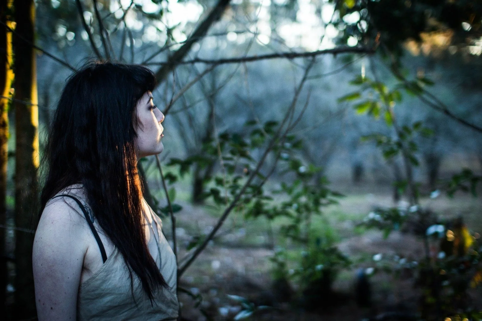 A young woman with long black hair and light skin standing outdoors in a forest, looking to the left, with trees and green foliage in the background during sunset.
