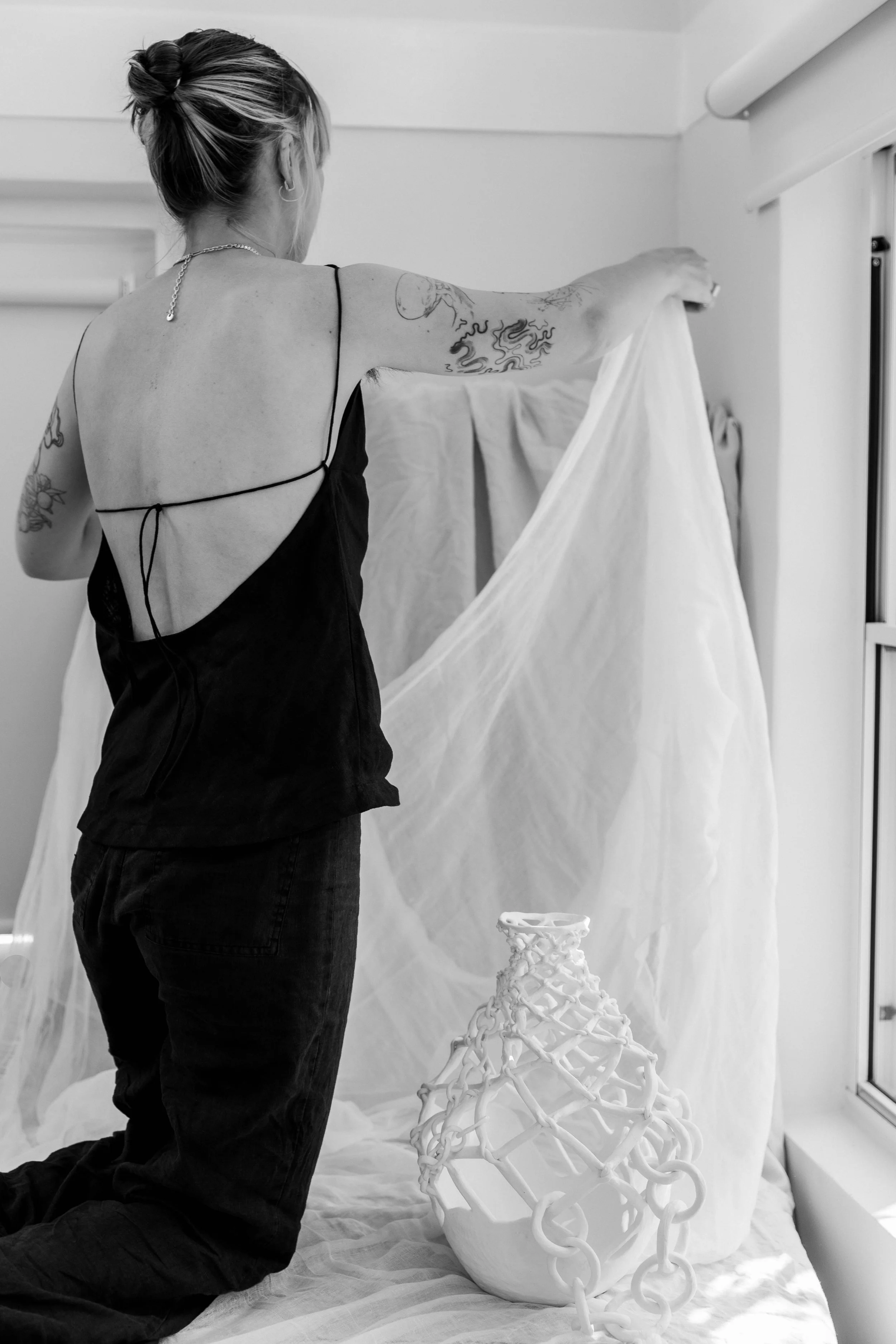 A woman with tattoos on her arms, wearing a black camisole, is arranging a sheer fabric in a room. There is a decorative ceramic vase with a lattice pattern on the floor and a window with blinds.