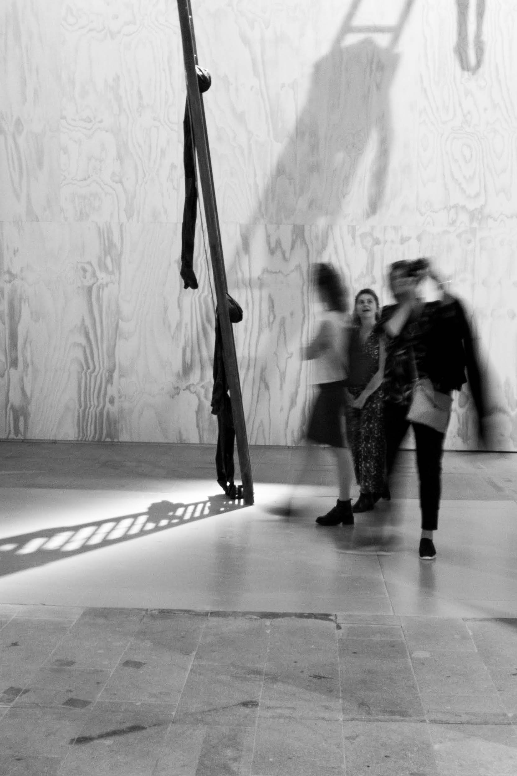 Three women walking past a wall with a shadow of a silicon figure hanging on a metal structure in an art gallery.