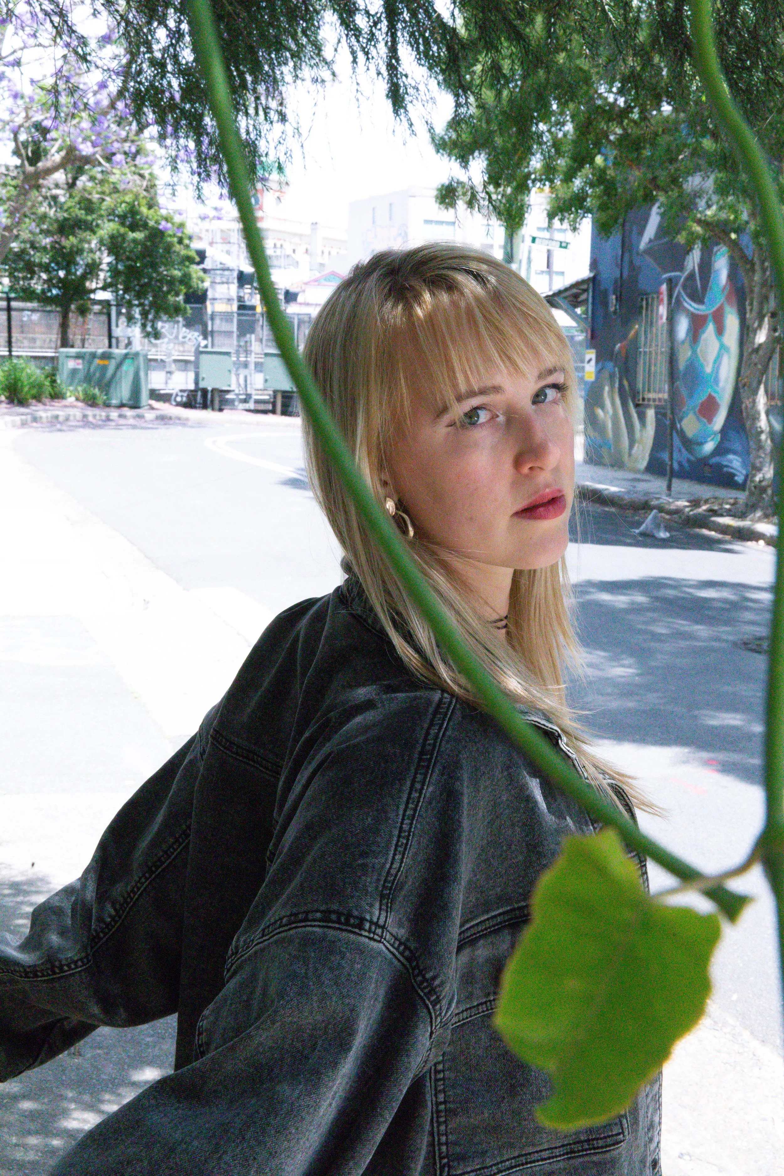 A woman with blonde hair and a black denim jacket standing outdoors in an urban setting, partially obscured by a large green leaf.