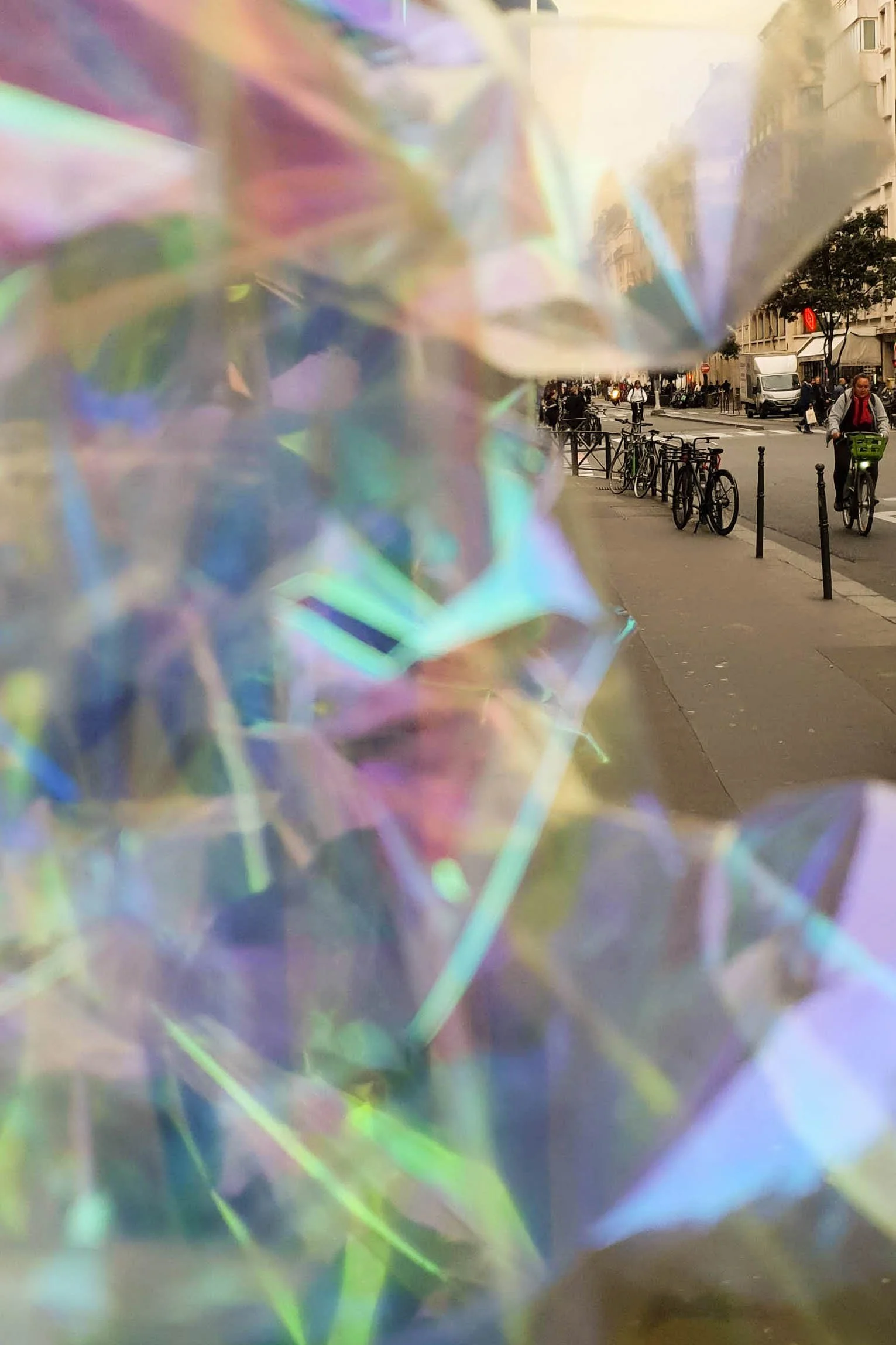 Close-up of iridescent, translucent materials with a city street scene in the background, including bicycles, pedestrians, buildings, and vehicles.