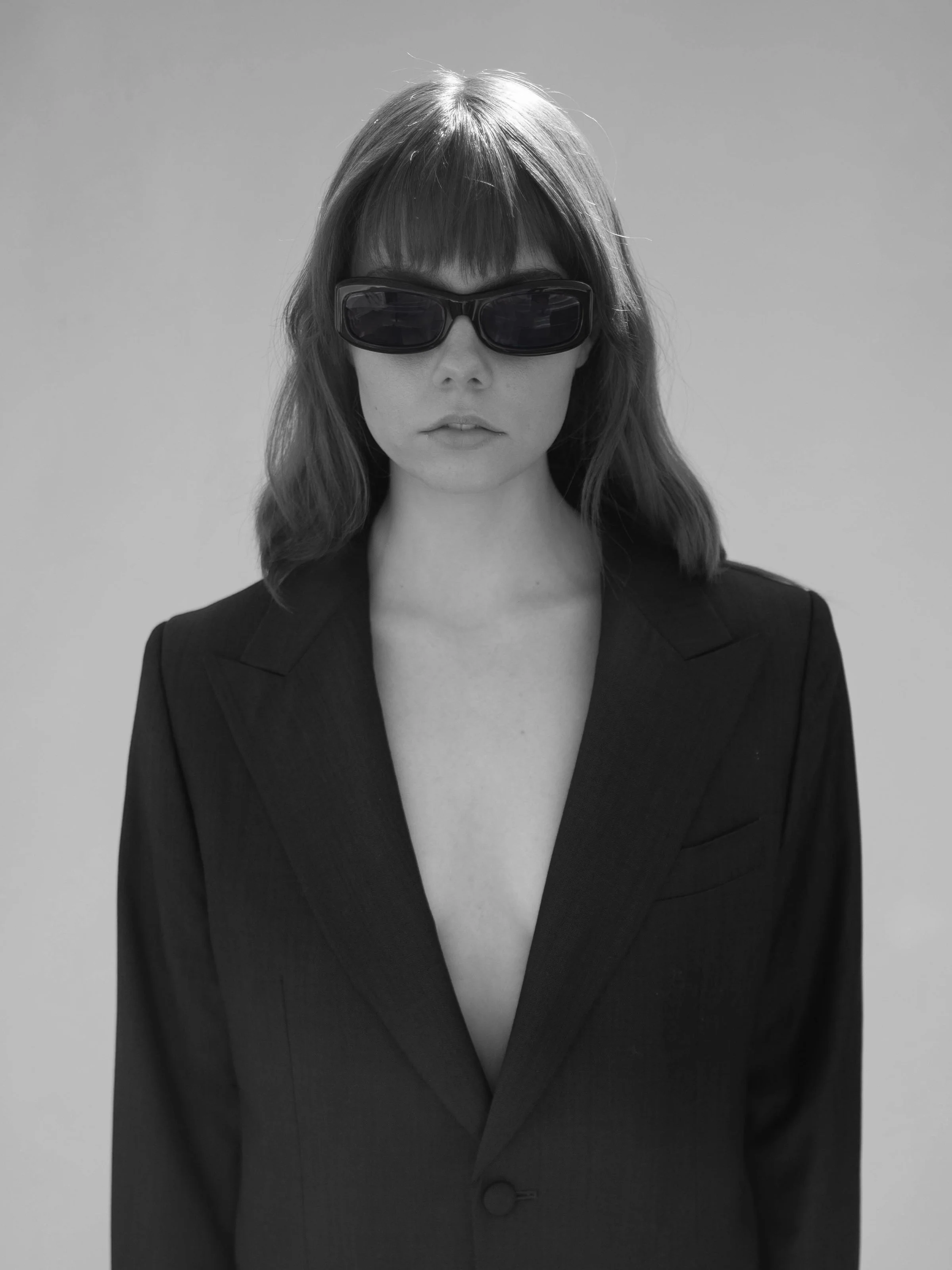 Black and white photo of a woman wearing dark sunglasses and a dark blazer with a deep V neckline, standing against a plain background.