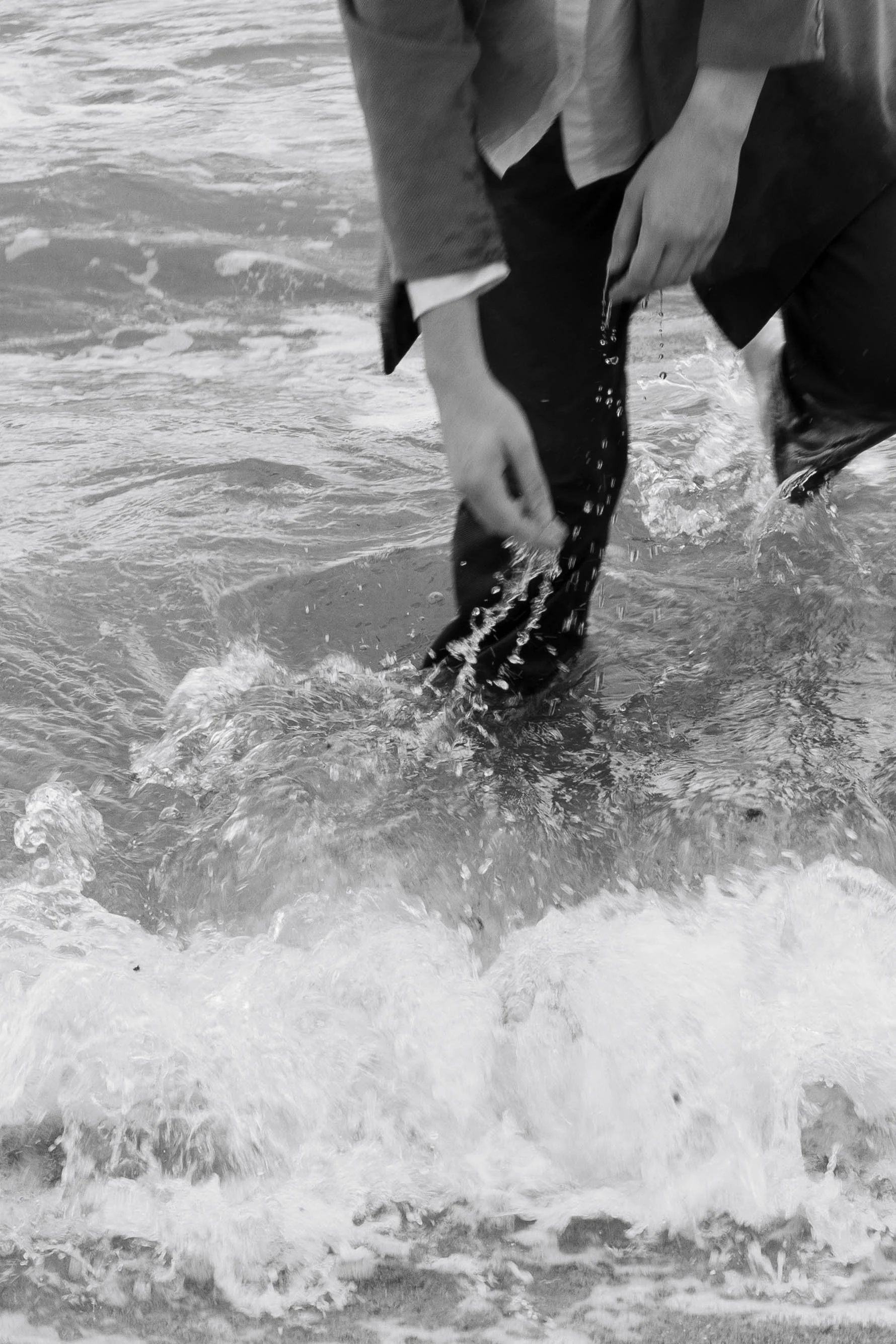 Person dressed in formal attire, including a blazer, crouching in shallow water at the beach.