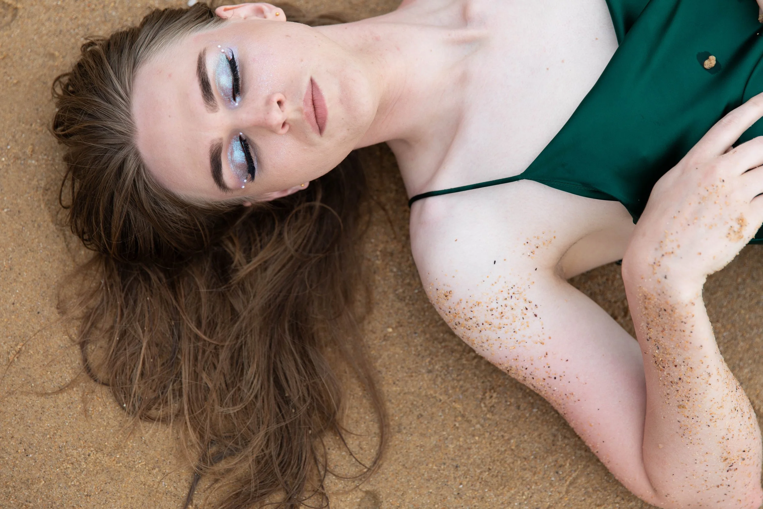 A woman with long brown hair lying on sandy ground, wearing a dark green dress, with sand on her arm and shoulder, and makeup with blue eyeshadow and lipstick.