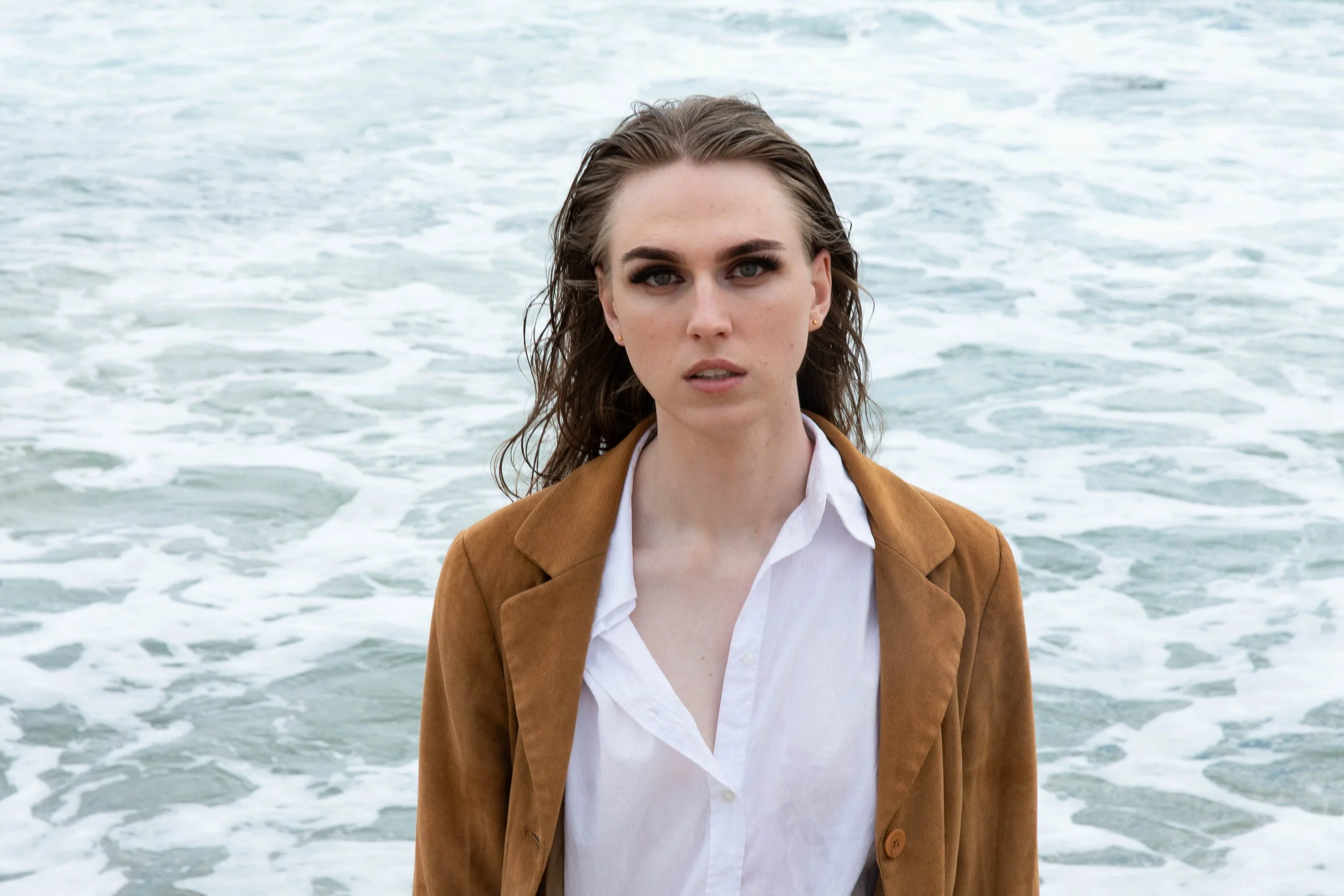 A woman with wavy brown hair and blue eyes standing in front of the ocean with waves crashing behind her.