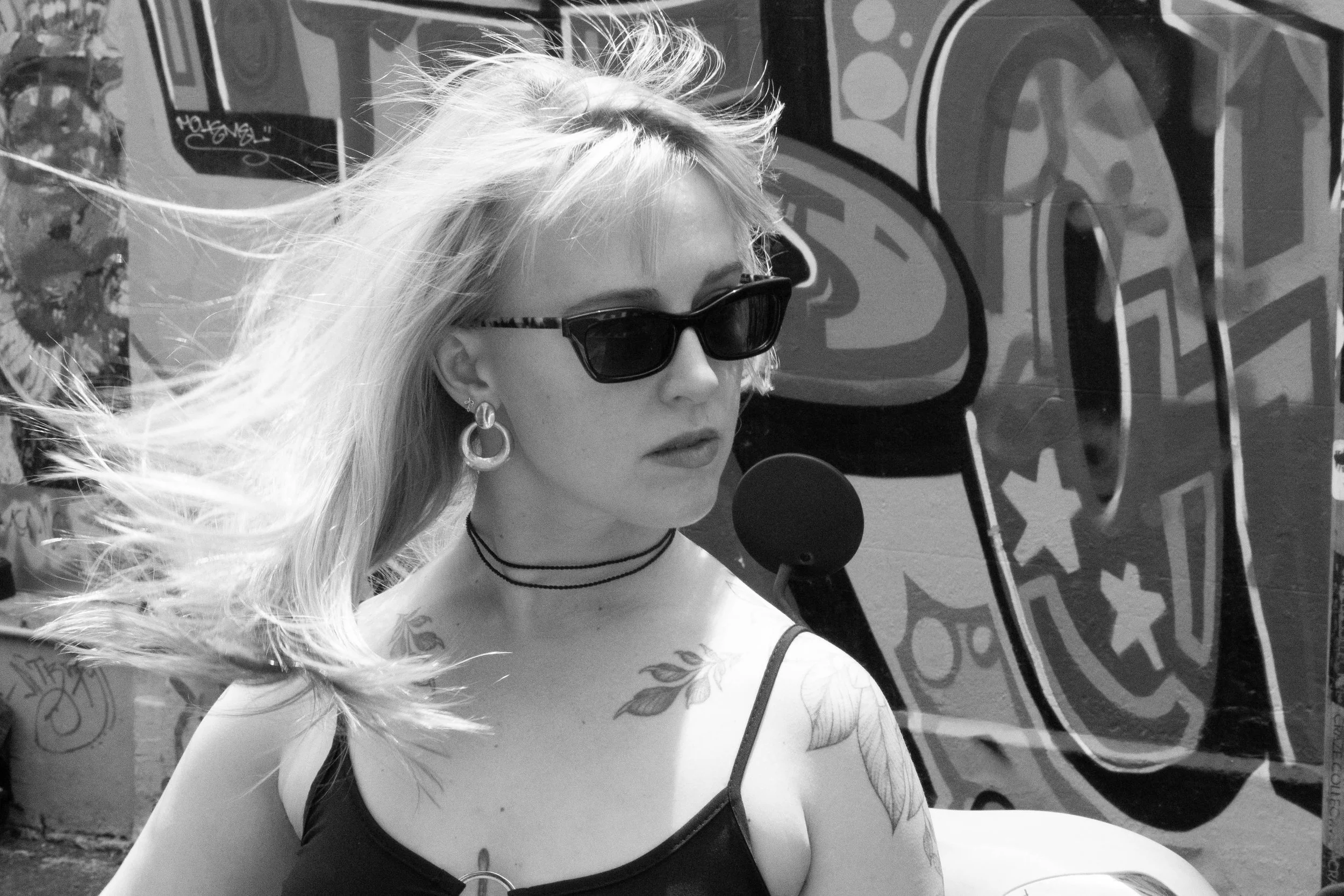 A woman with blonde hair wearing sunglasses and hoop earrings pictured in front of graffiti art.
