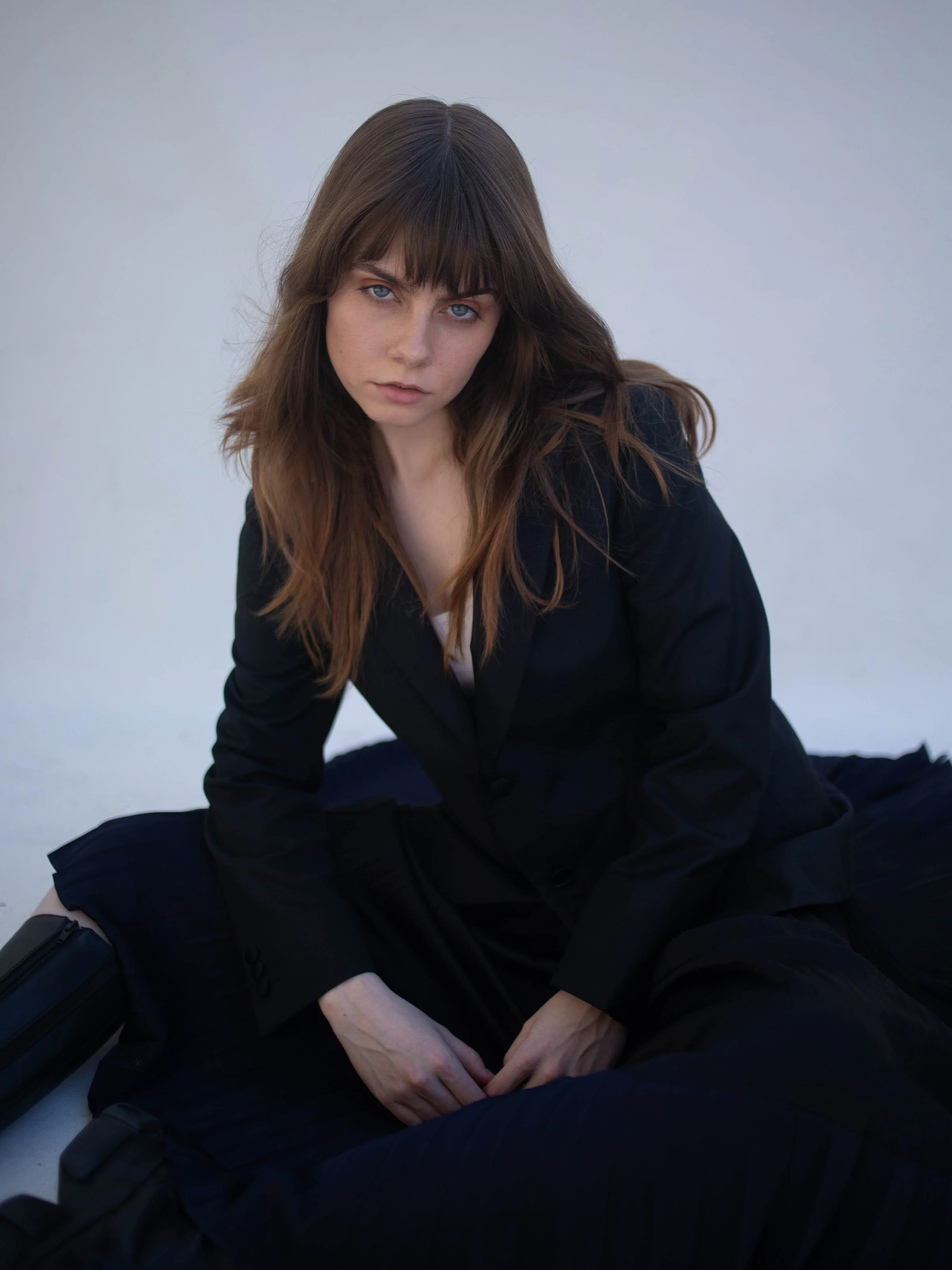 A young woman with long hair and bangs, wearing a black blazer and pants, sits on the ground against a plain light background, looking directly at the camera.