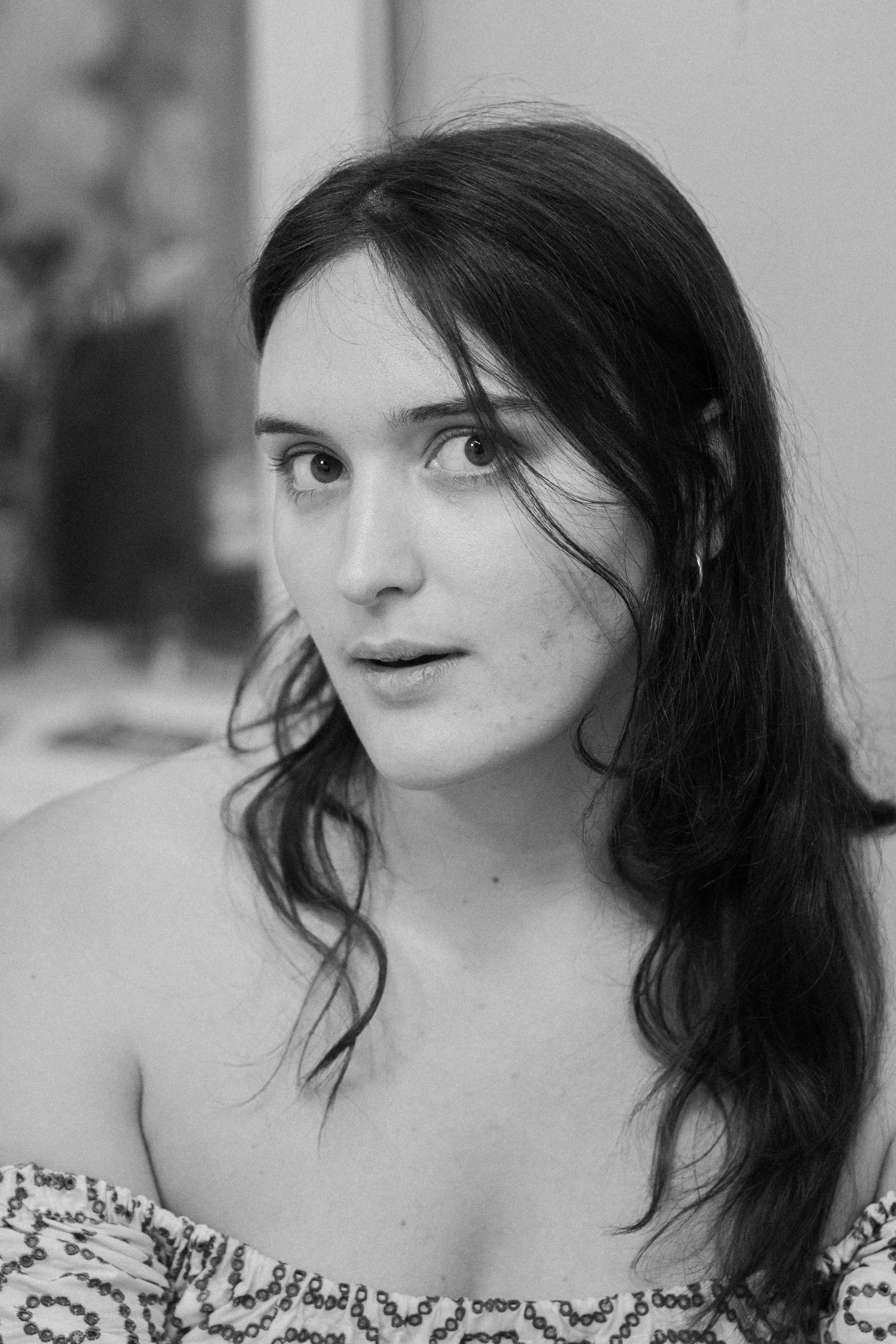 Black and white close-up portrait of a young woman with long dark hair, looking directly at the camera with a slightly open mouth. She is wearing an off-the-shoulder top with a patterned design.