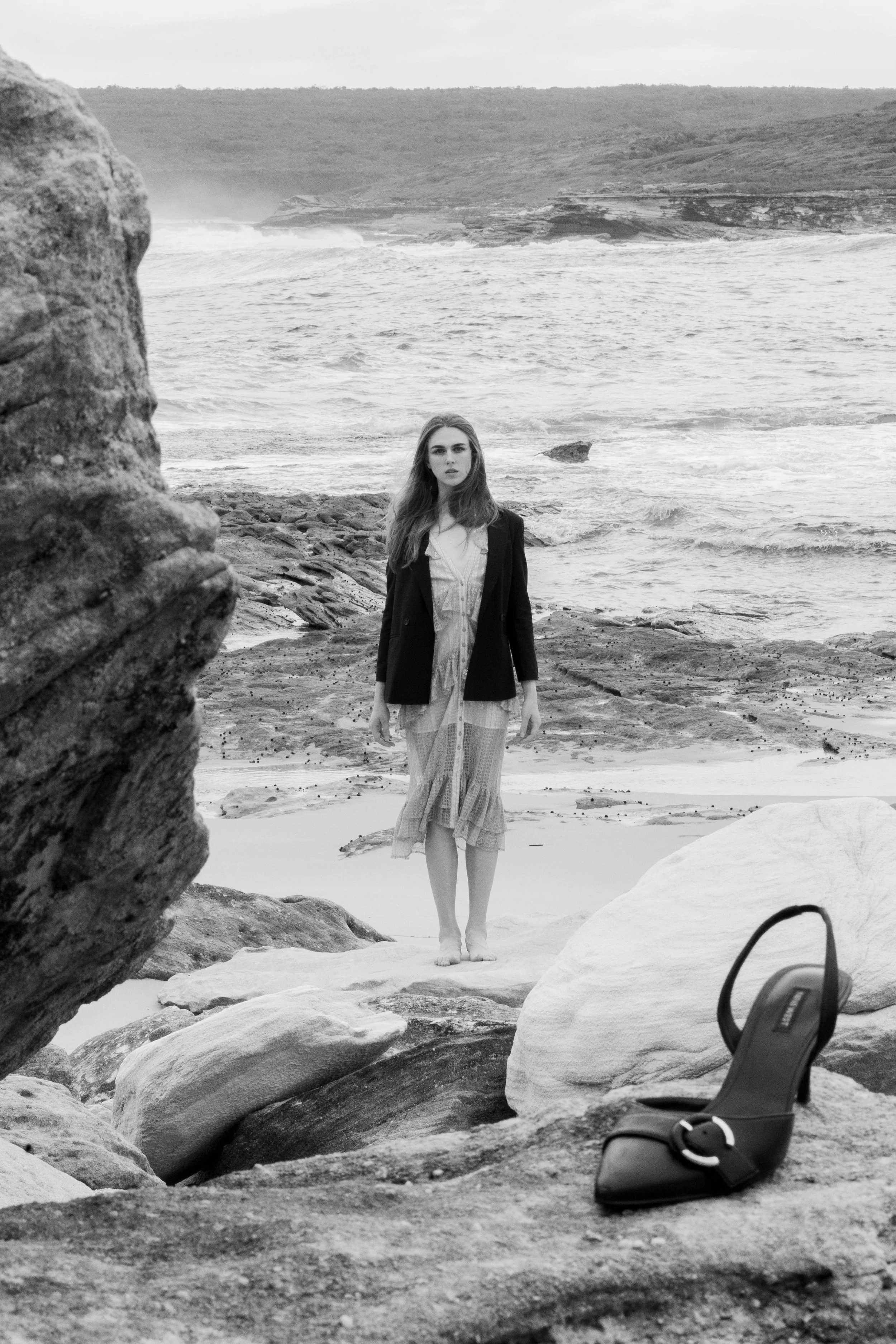 A woman in a dress and blazer standing barefoot on rocks at a beach, with a high heel shoe in the foreground.