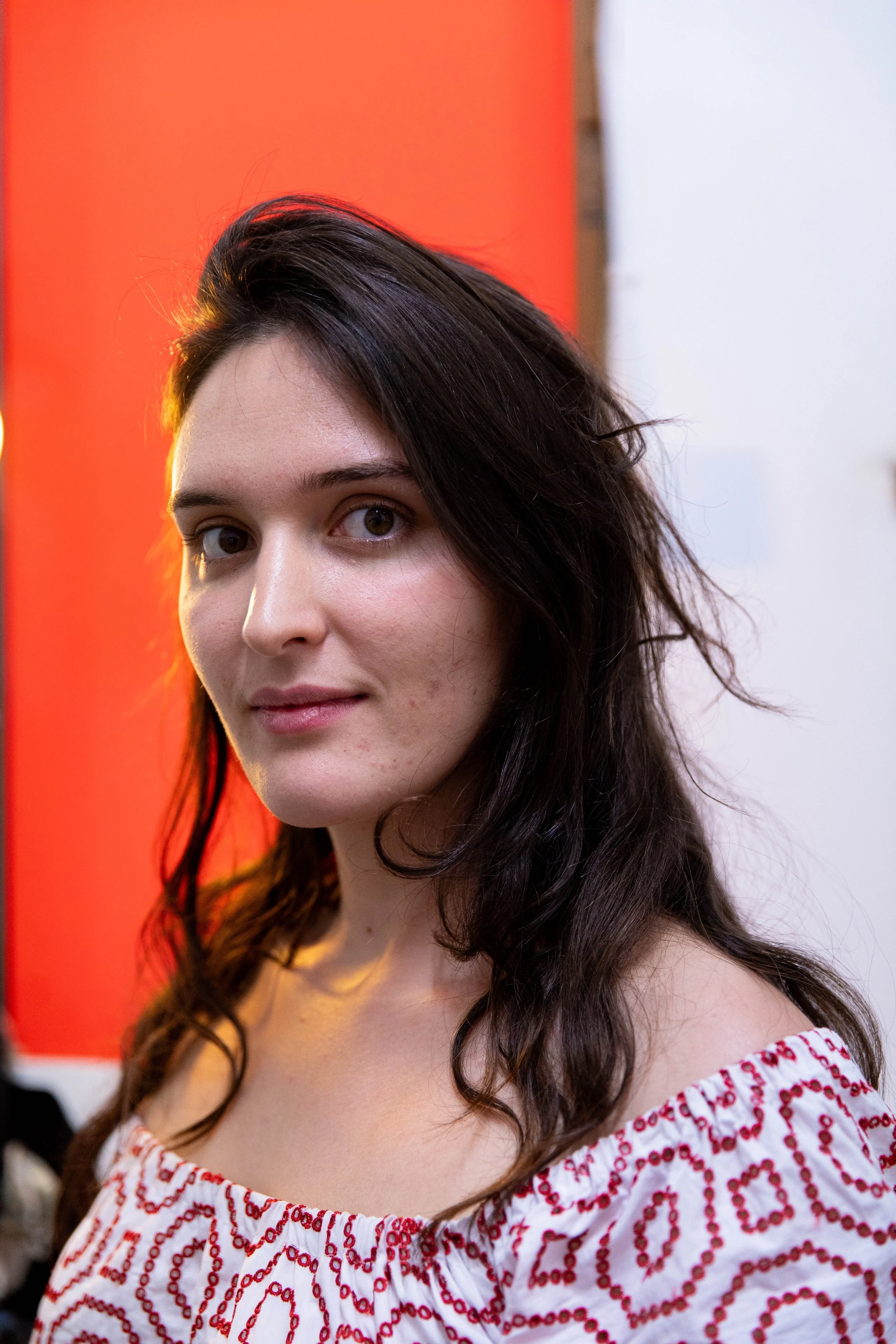A portrait of a young woman with dark brown hair and hazel eyes, wearing am off-shoulder top with red, standing indoors in front of a bright red wall, illuminated by warm lighting.