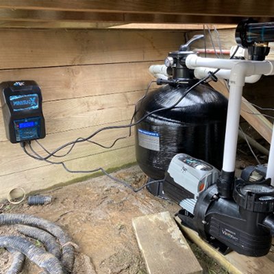 Pool pump and filter system setup in a wooden enclosure, including black filter tank, white PVC pipes, and electronic control panel with wires.