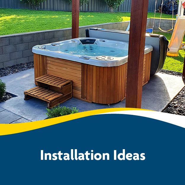 Outdoor hot tub with wooden paneling and steps, set on a concrete patio, surrounded by green grass and a stone wall.