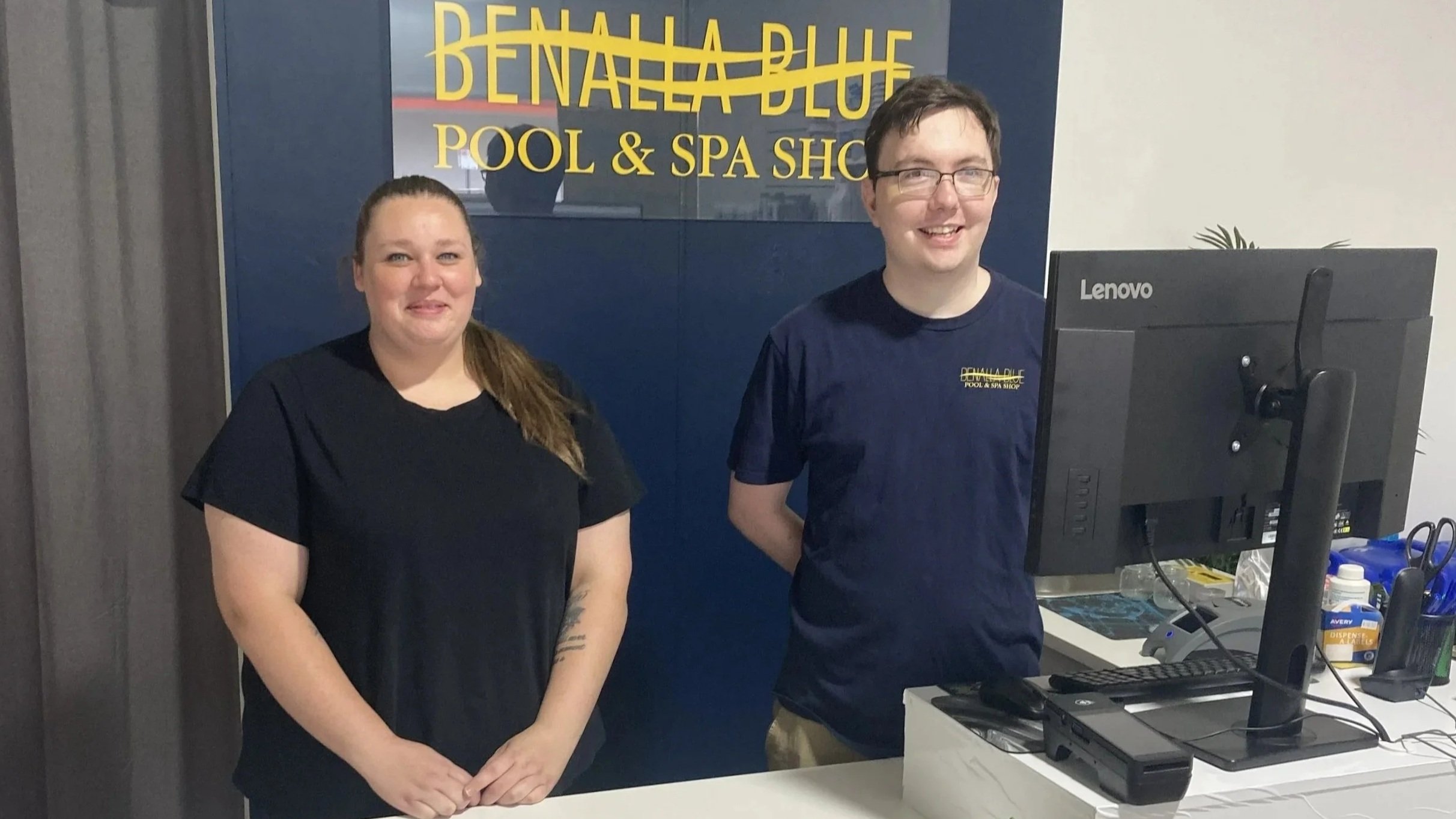 Two employees at the reception desk of Benalla Blue Pool & Spa. The woman on the left has long brown hair and is wearing a black shirt. The man on the right has short dark hair, glasses, and is wearing a navy blue shirt with the business logo. Behind them is a sign that reads 'Benalla Blue Pool & Spa' and a computer monitor is on the desk.