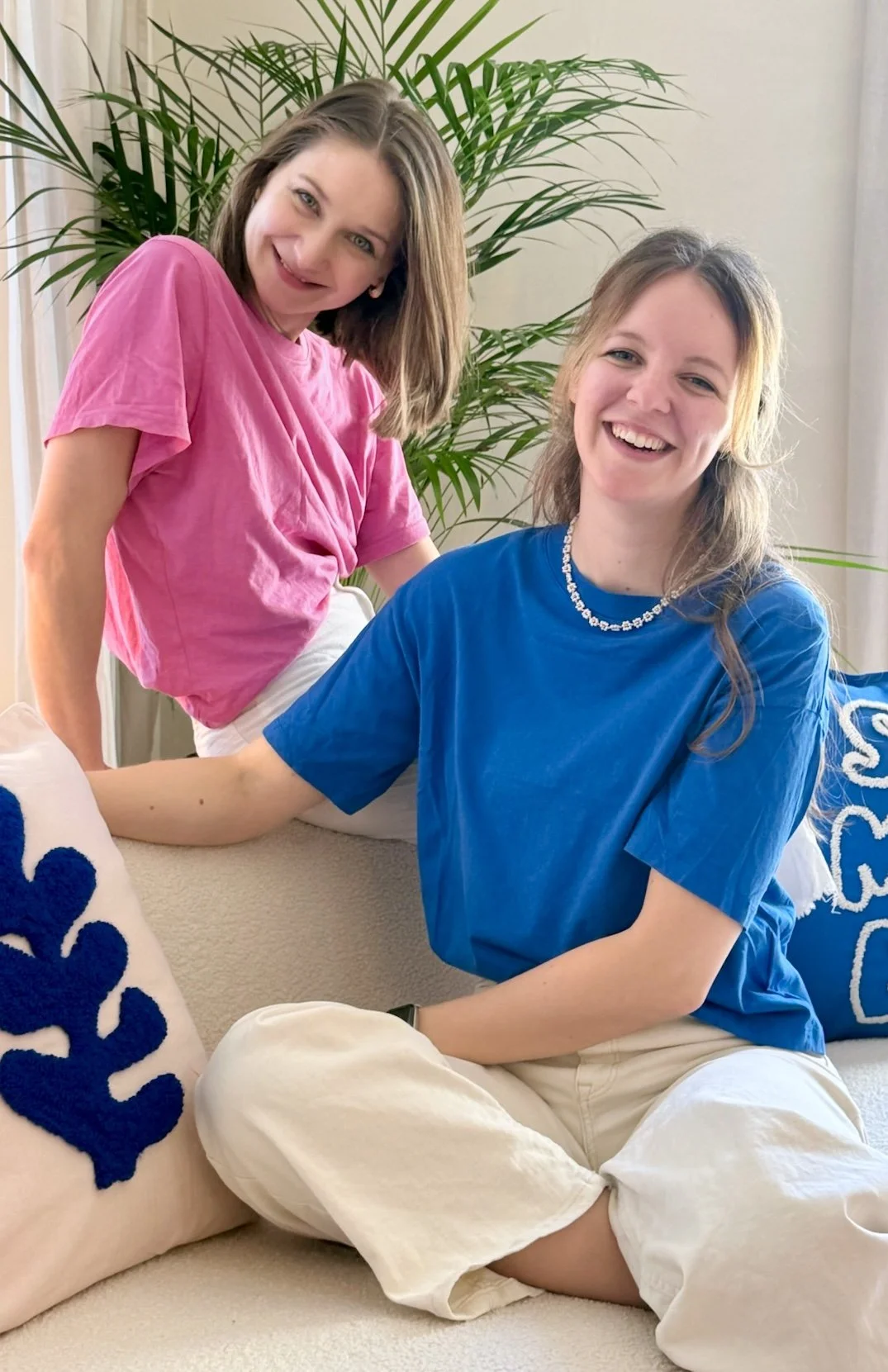 Zwei lachende Frauen in einem Wohnzimmer, eine sitzt auf einem Sofa in einem blauen T-Shirt und die andere steht dahinter in einem pinken T-Shirt. Es gibt grüne Pflanzen im Hintergrund und Kissen mit blauen Mustern auf dem Sofa.