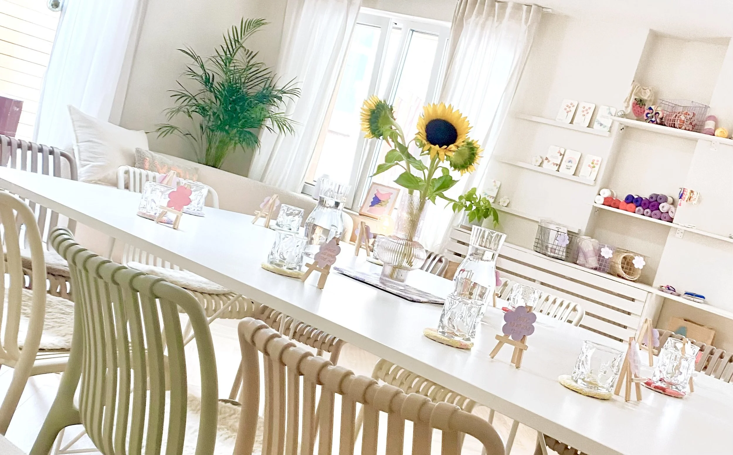 Ein hell erleuchteter Raum mit weißem Esstisch, dekoriert mit einem großen Blumenstrauß aus Sonnenblumen in einer Glasvase, kleineren Glasvasen und Platzkarten. Es gibt Stühle aus Holz mit weißen Kissen. Im Hintergrund sind weiße Regale mit Handarbei