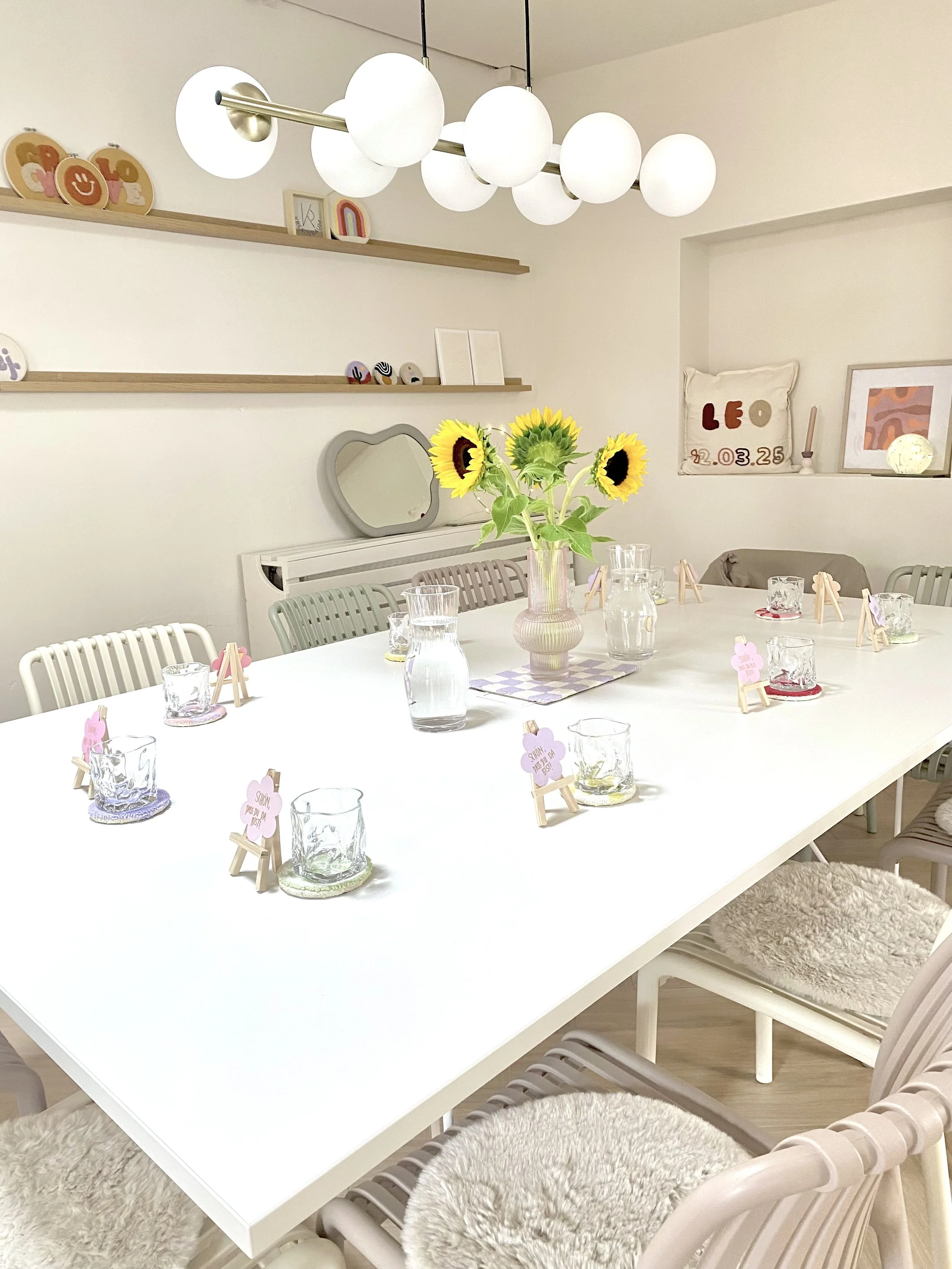 Ein weißer Esstisch mit einem Blumenstrauß aus Sonnenblumen in der Mitte, umgeben von Gläsern, kleinen Untersetzern und Mini-Wägelchen mit pinken und lila Papierschilder.