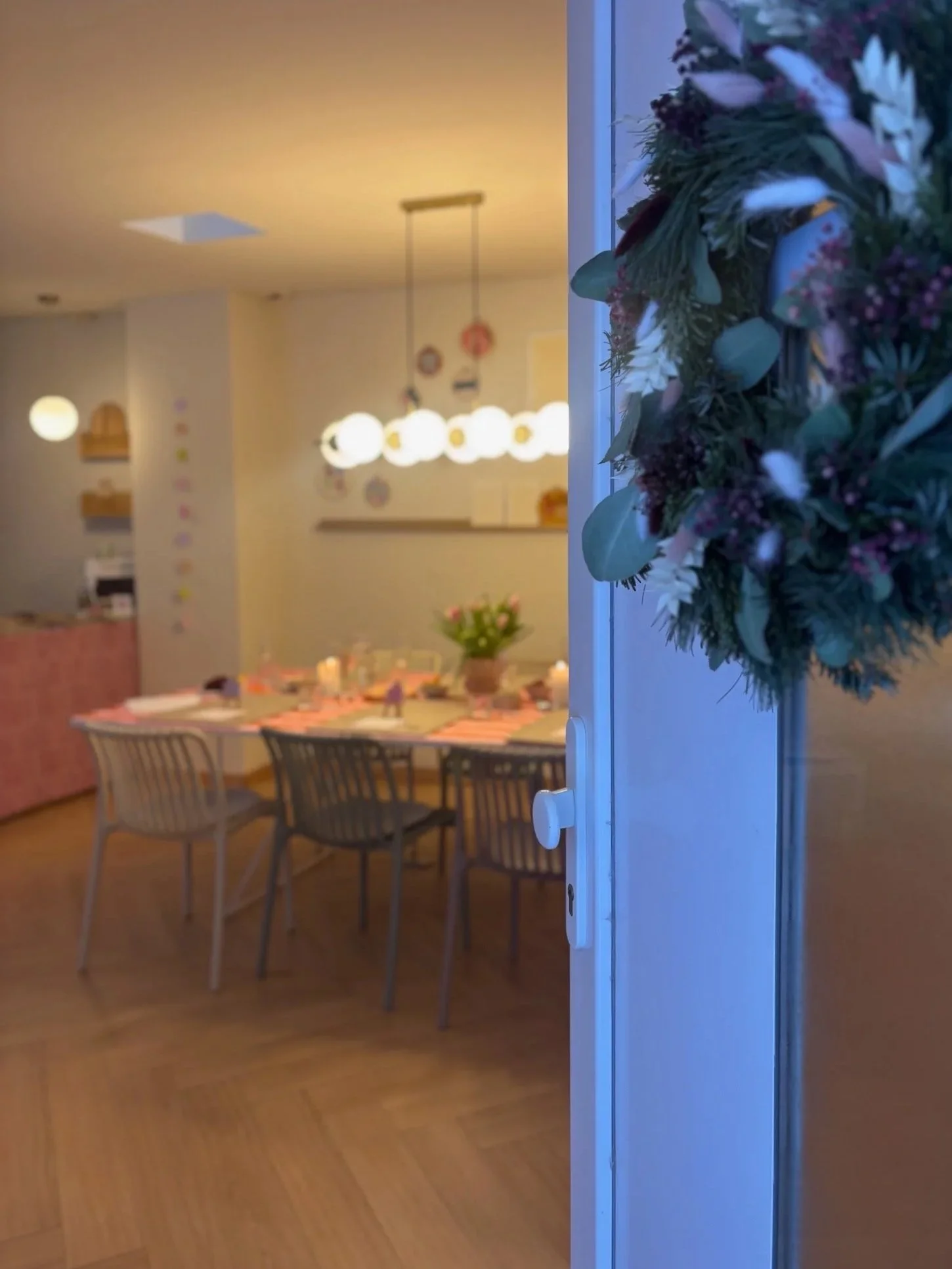 Blick durch eine Tür auf einen festlich gedeckten Esstisch mit Blumen, Kerzen und gedecktem Geschirr in einem Wohnzimmer mit moderner Beleuchtung.