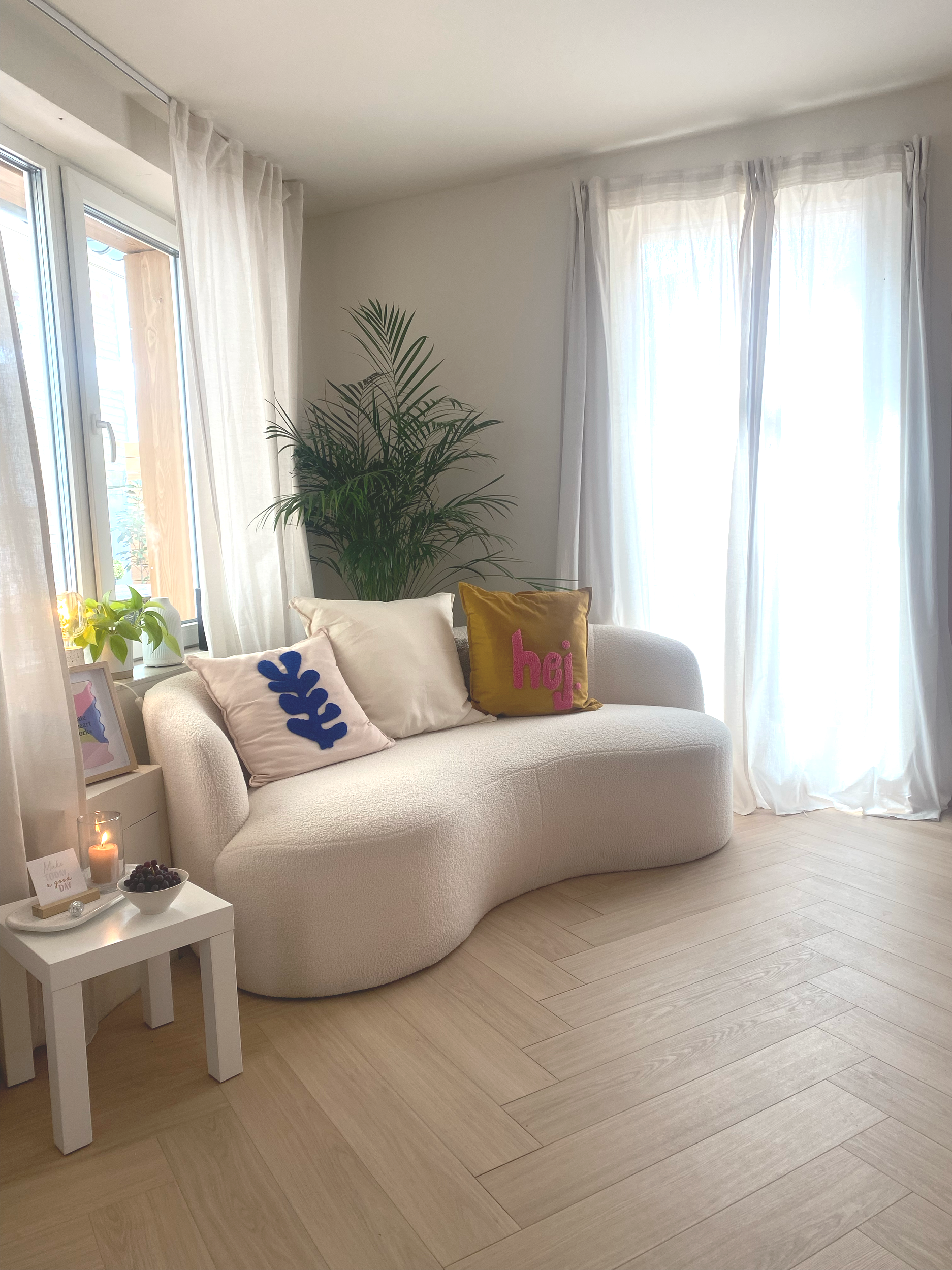 Gemütliches Wohnzimmer mit weißen Sofa, bunten Kissen, Pflanze, Seiten Tisch mit Kerze und Obst, große Fenster mit weißen Vorhängen.