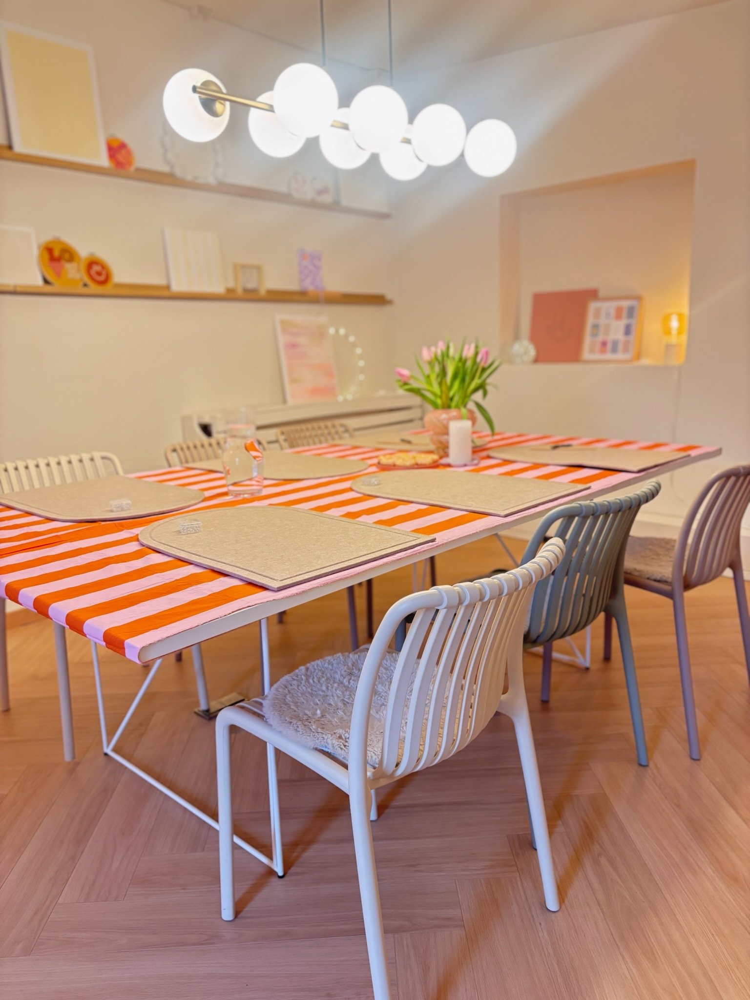 Ein heller Esstisch mit orange-weiß gestreifter Tischdecke, mit vier beige Platzsets, eine Vase mit Tulpen, eine Kerze und ein Glas Wasser. Im Hintergrund sind weiße Wände mit Regalen mit Dekorationsartikeln und Kunstwerken, eine moderne Hängelampe m