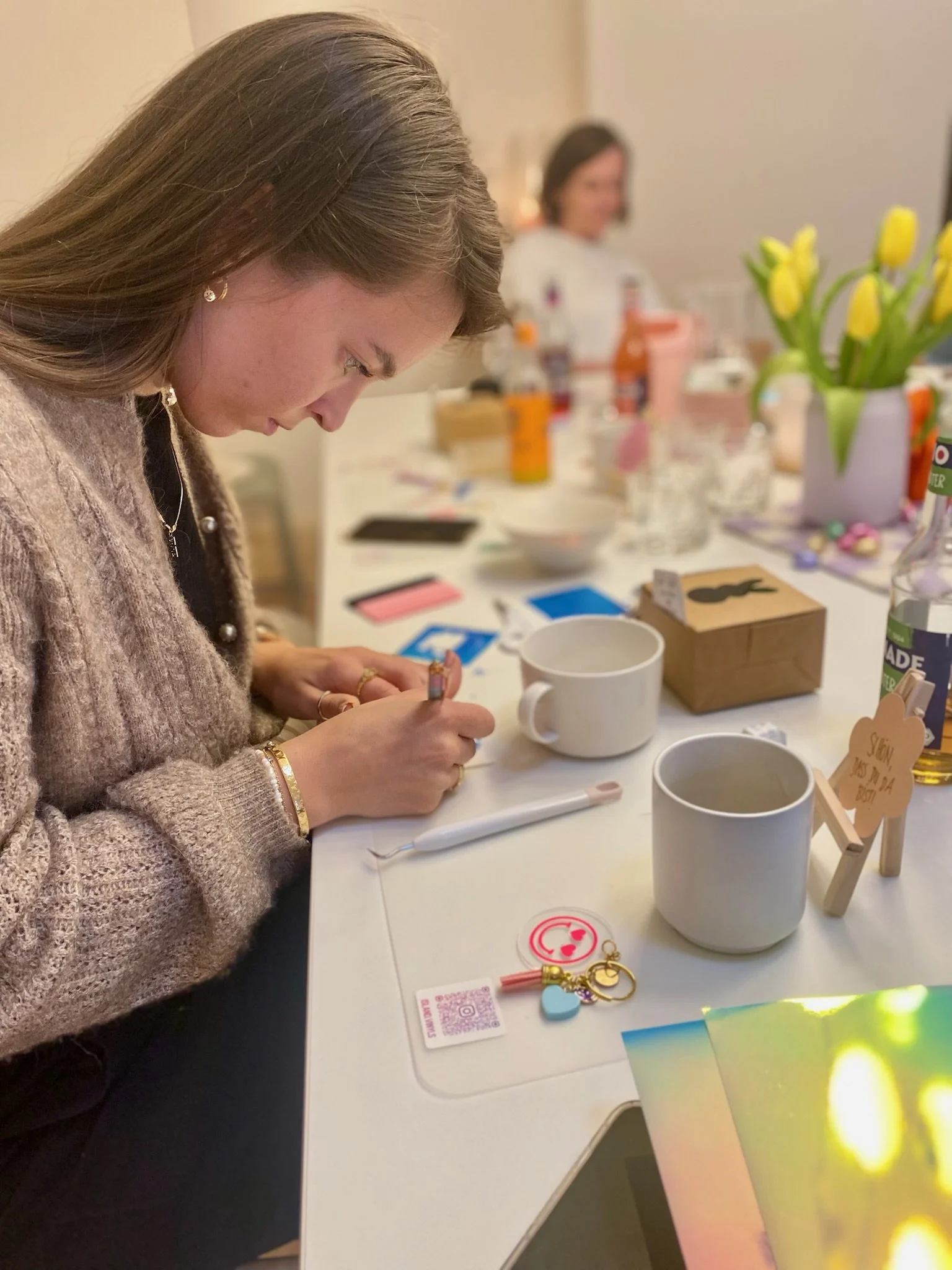 Eine junge Frau mit braunen Haaren sitzt an einem Tisch bei einem kreativen Workshops, im Hintergrund eine andere Person ebenfalls am Tisch sitzt.
