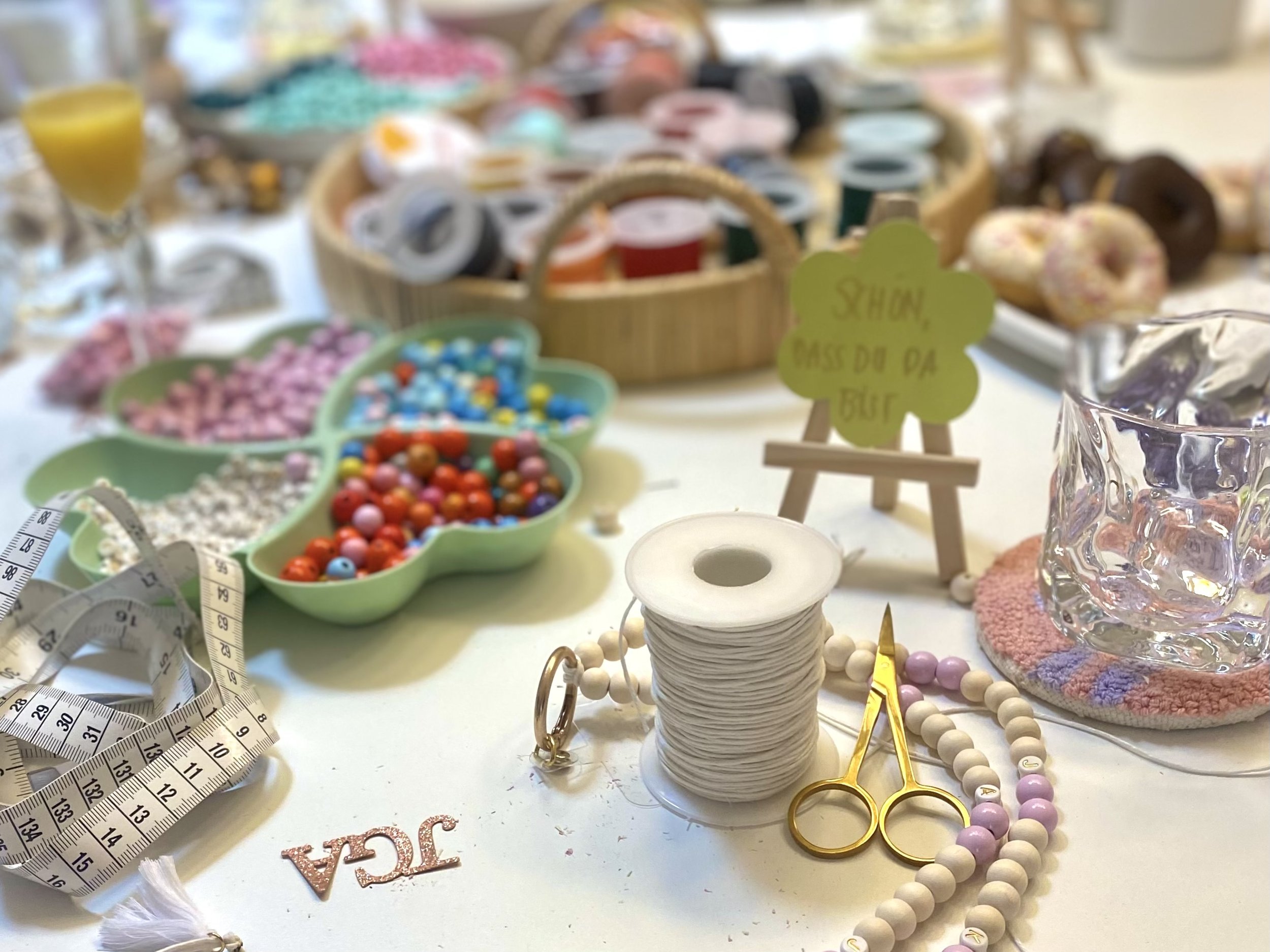 Bunte Perlen, Bastelmaterialien, ein Schriftband, eine Glasvase, Donuts und ein Körbchen mit buntem Garn auf einem Basteltisch