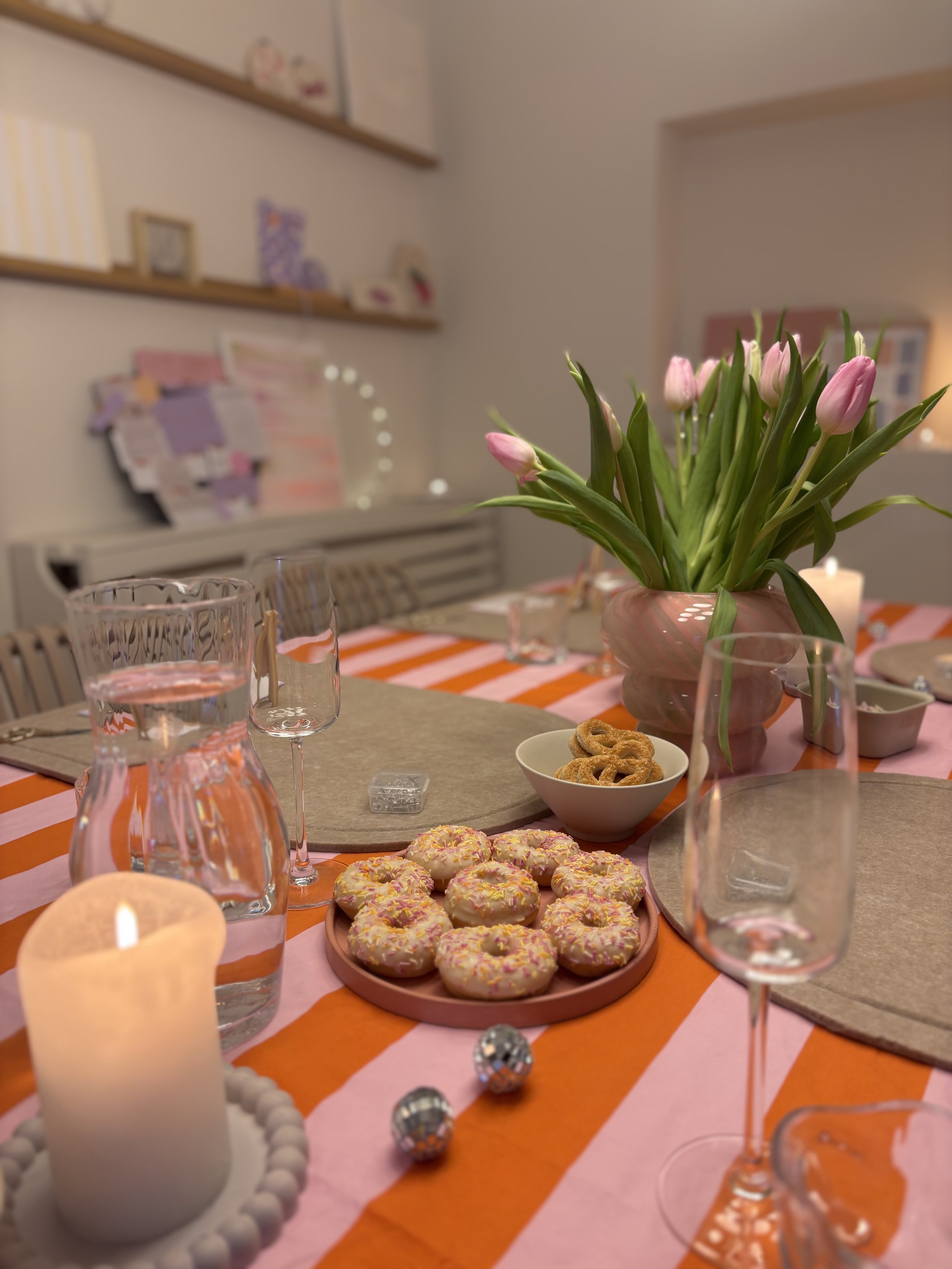 Der Tisch ist für eine Party oder Feier gedeckt, mit einem Blumenstrauß mit pinken Tulpen, einem Kerzenhalter mit einer LED-Kerze, Gläsern, einer Platte mit Donuts mit bunten Streuseln, einer Schüssel mit Keksen und einem weiteren kleinen Teller. Es 