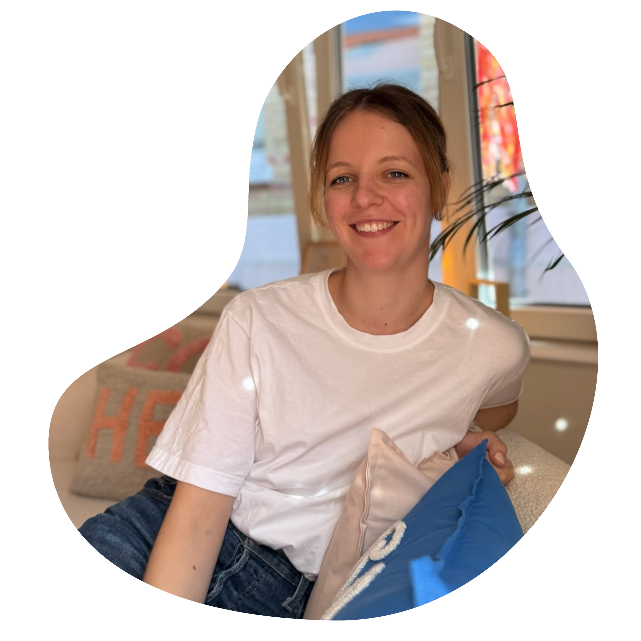 Eine junge Frau sitzt auf einem Sofa vor einem Fenster, lächelt und trägt ein weißes T-Shirt. Im Hintergrund sind Kissen und eine Pflanze sichtbar.