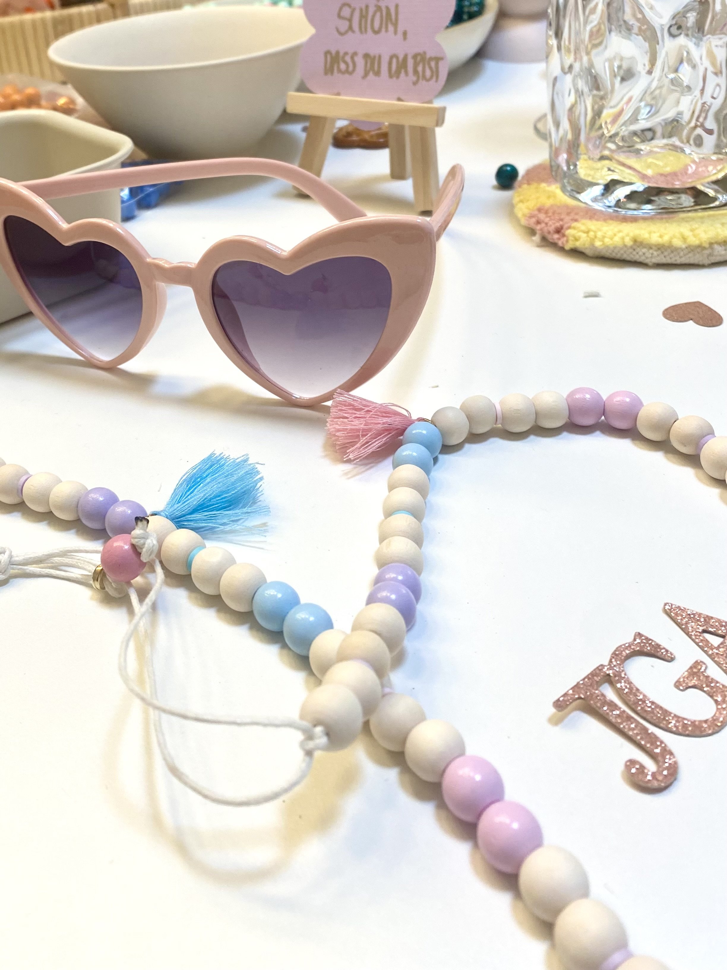 Bunte Perlenketten, eine rosa Herzbrille mit lila Sonnenbrillengläsern, Cupcakes und Dekorationen auf einem Festtisch.