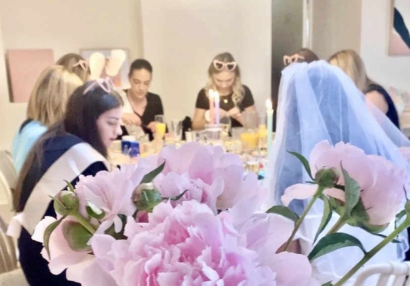 Menschen feiern an einem Tisch, Blumen im Vordergrund, jemand trägt eine Hochzeitstracht und einem Schleier.