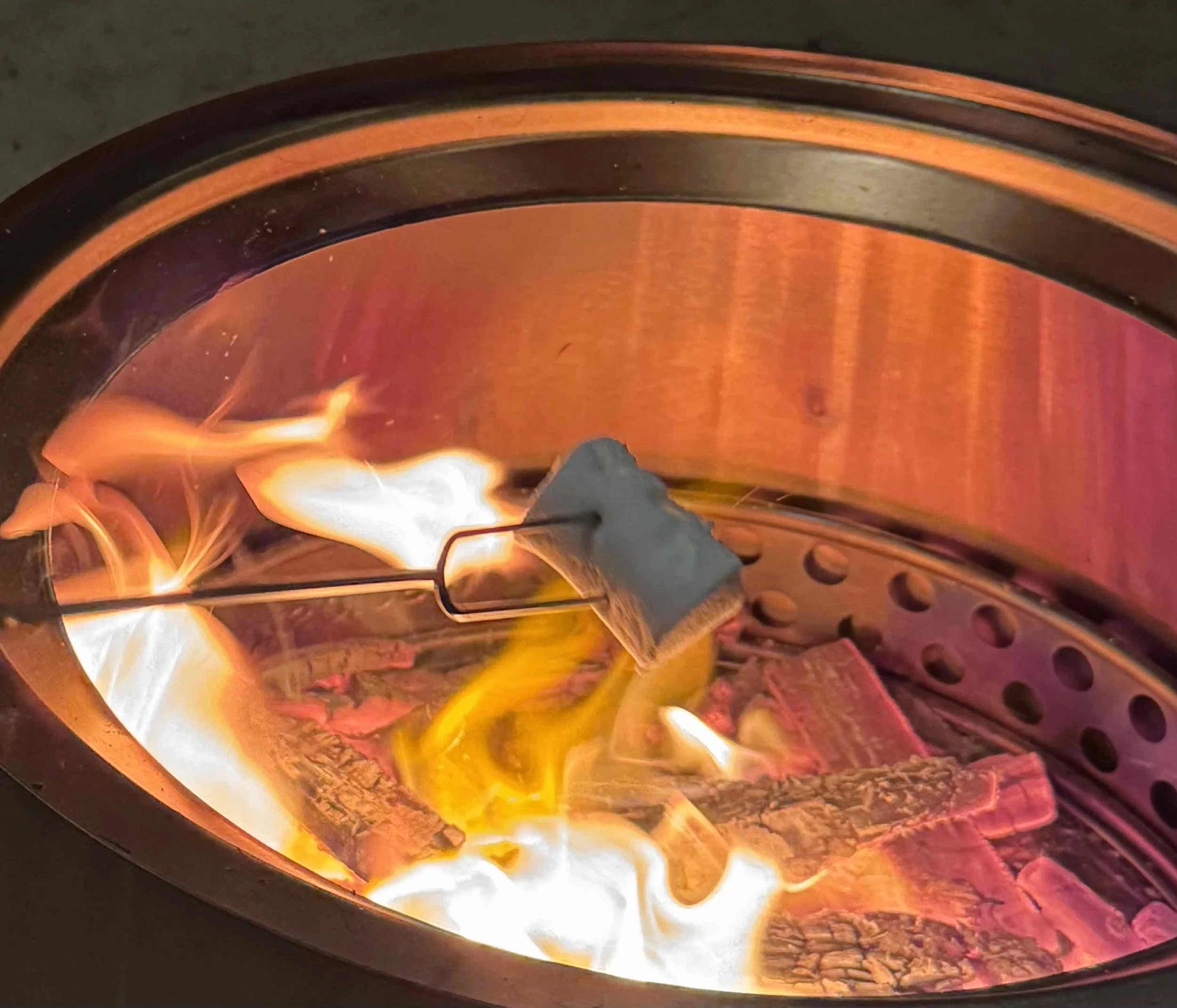 A close-up of a fire inside a wood stove, with flames burning around pieces of wood and a metal rod holding a marshmallow over the flames.