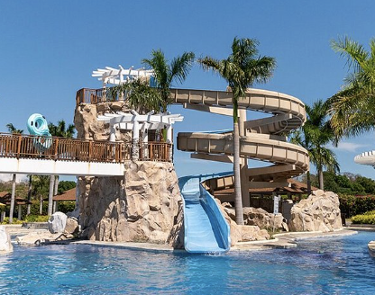 A water park with a large water slide, rock formations, palm trees, and a pool.