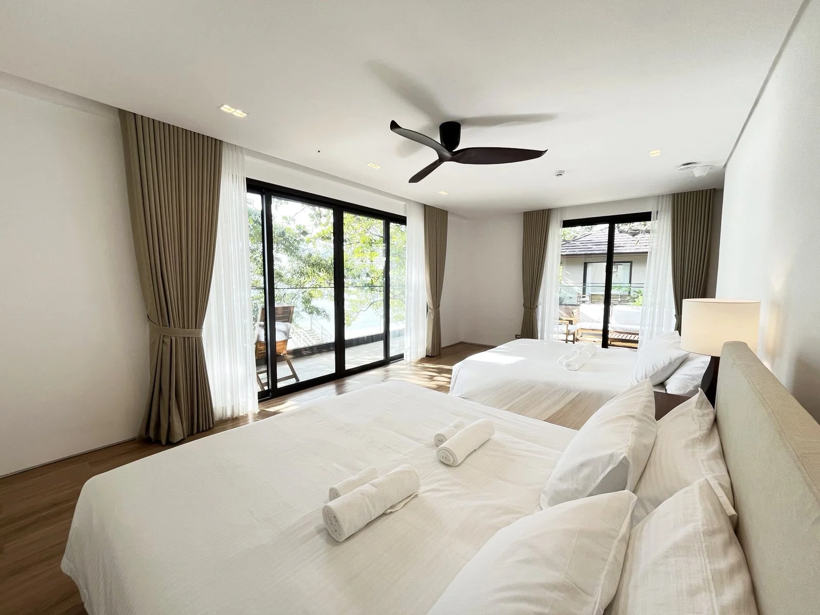 A bright, modern hotel room featuring two large beds with white linens and rolled towels, a bedside lamp, ceiling fan, and a balcony with outdoor furniture accessible through sliding glass doors, overlooking trees and neighboring buildings.