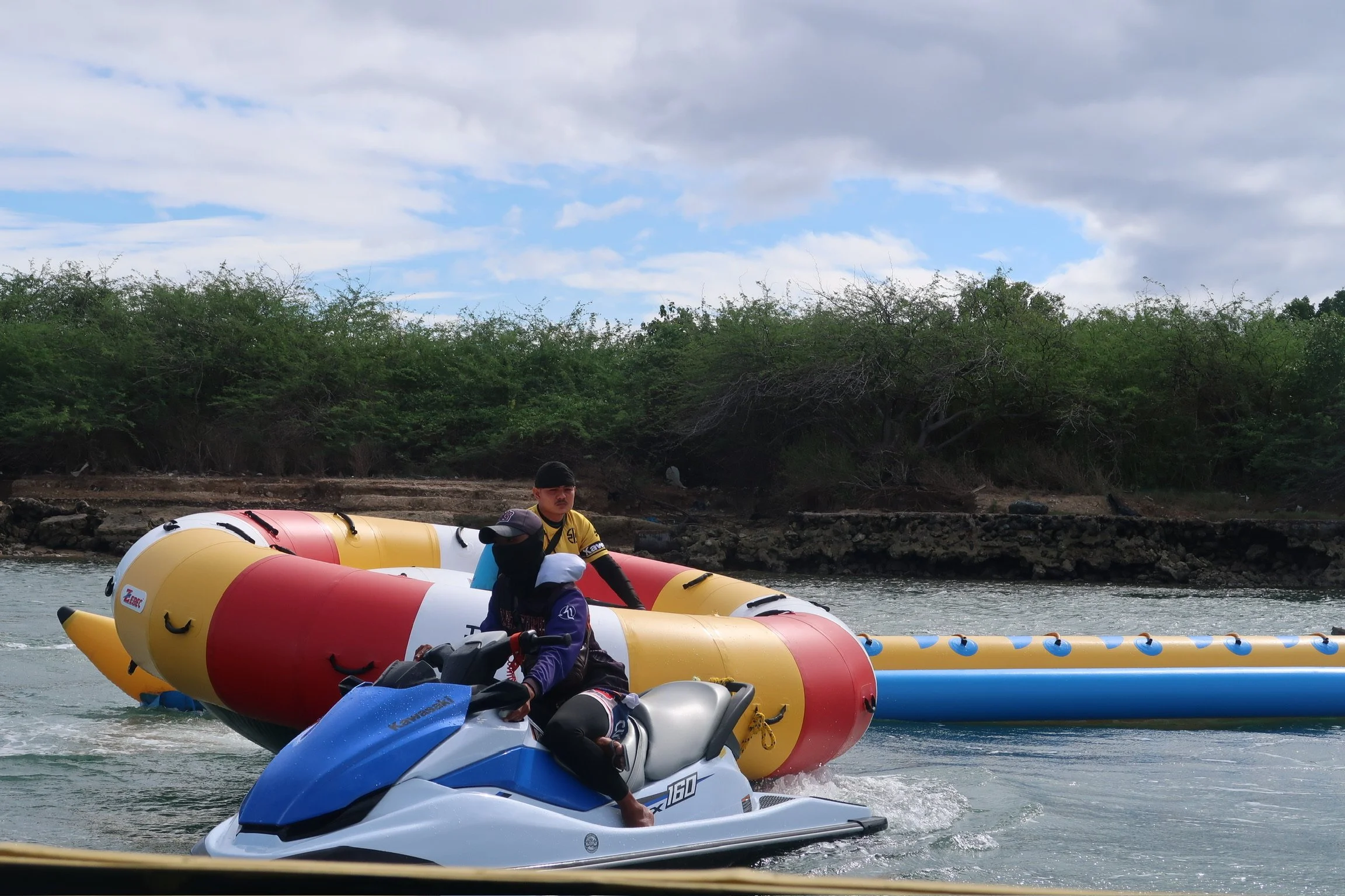 Water sports: people riding a jet ski beside a large inflatable raft on a river (banana boat), with trees and beautiful views of trees and the ocean in the background.