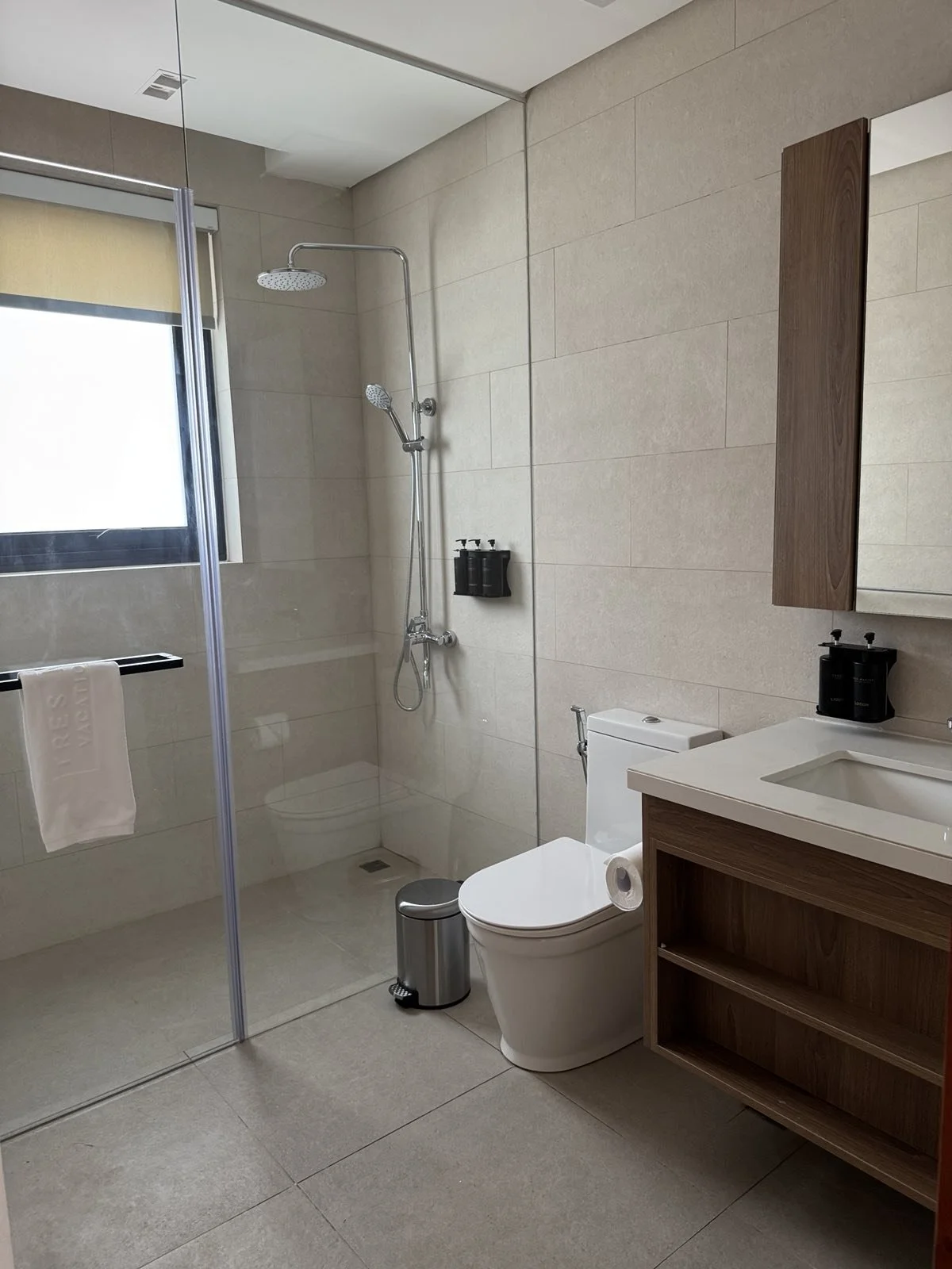 Modern bathroom with a shower area, toilet, and wooden vanity with a white sink. There is a window with a blind and a towel hanging on a shelf. The shower has a rain head and handheld showerhead, with toiletries mounted on the wall.
