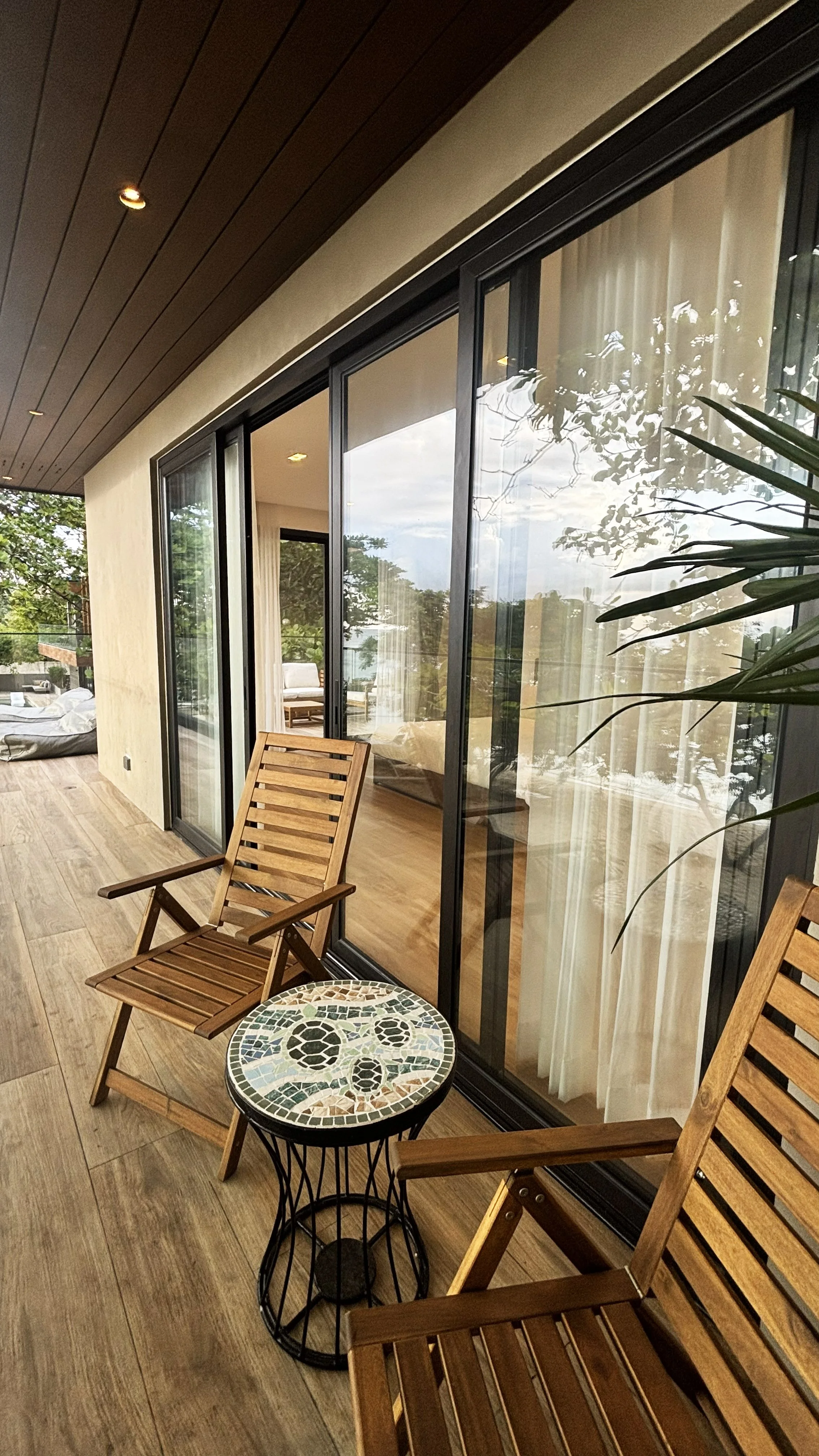 A cozy outdoor balcony with wooden chairs, a small mosaic-topped table, glass sliding doors, and greenery outside.