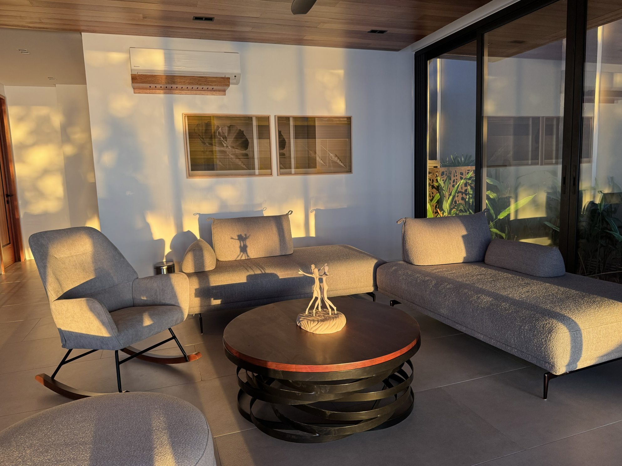 Living room with beige sofas, a rocking chair, and a black round coffee table with a sculpture on top. Sunlight casts shadows on the white wall and through large glass sliding doors leading to greenery outside. Wooden ceiling paneling and framed artw