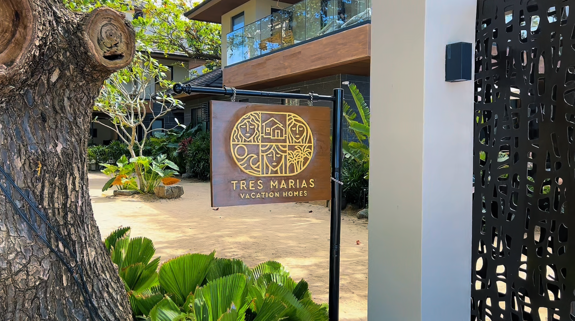 Wooden sign for Tres Marias Vacation Homes with a decorative logo featuring stylized faces and natural elements, located near a large tree with green foliage, black decorative fencing, and a modern building in the background.