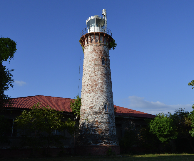 A tall old brick lighthouse with a glass observation deck on top, attached to a building with a red roof, surrounded by trees under a clear blue sky.