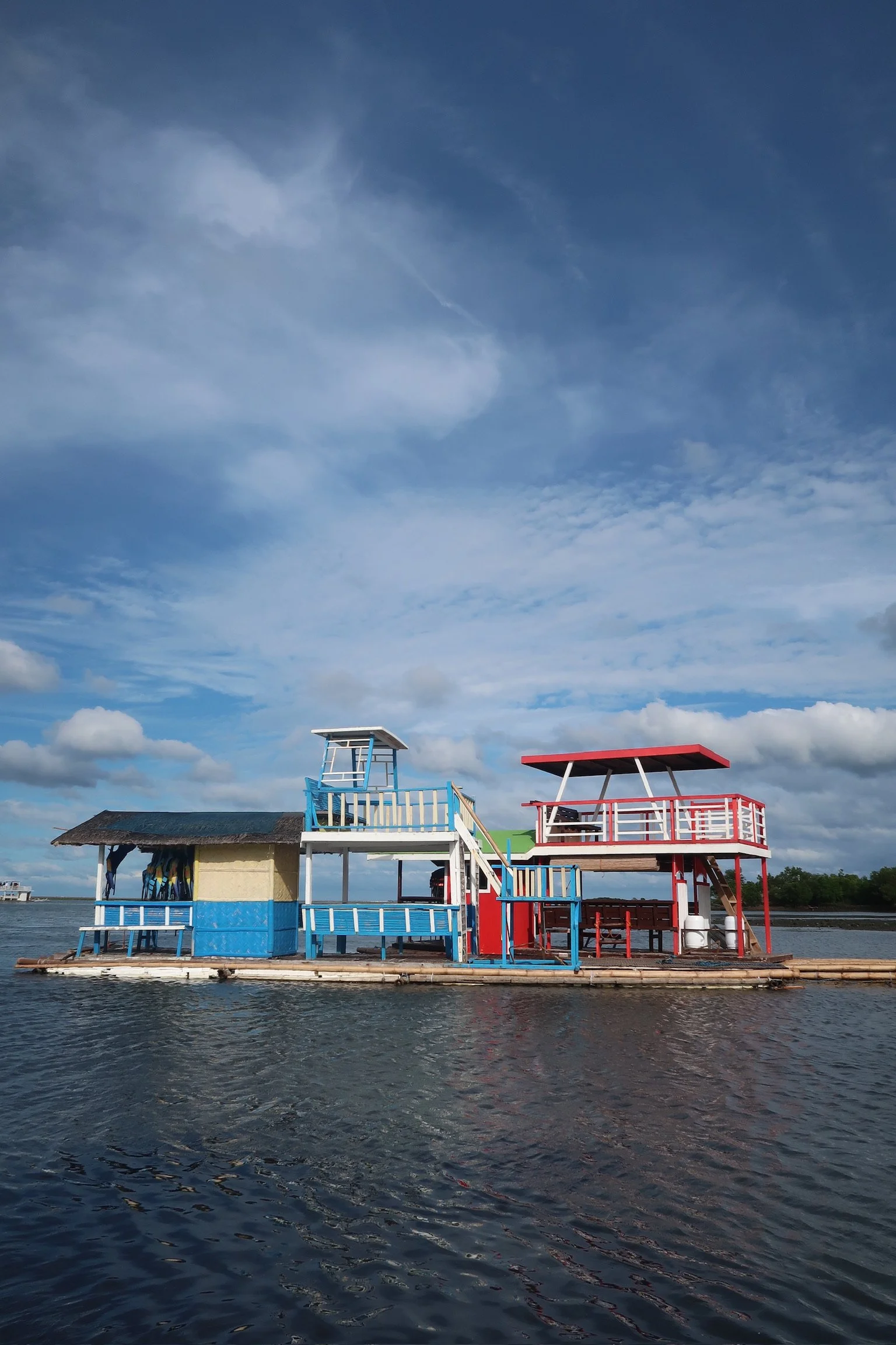 Colorful floating structure on water, resembling a lakeside or seaside bar or restaurant with boat-like features, under a partly cloudy sky.