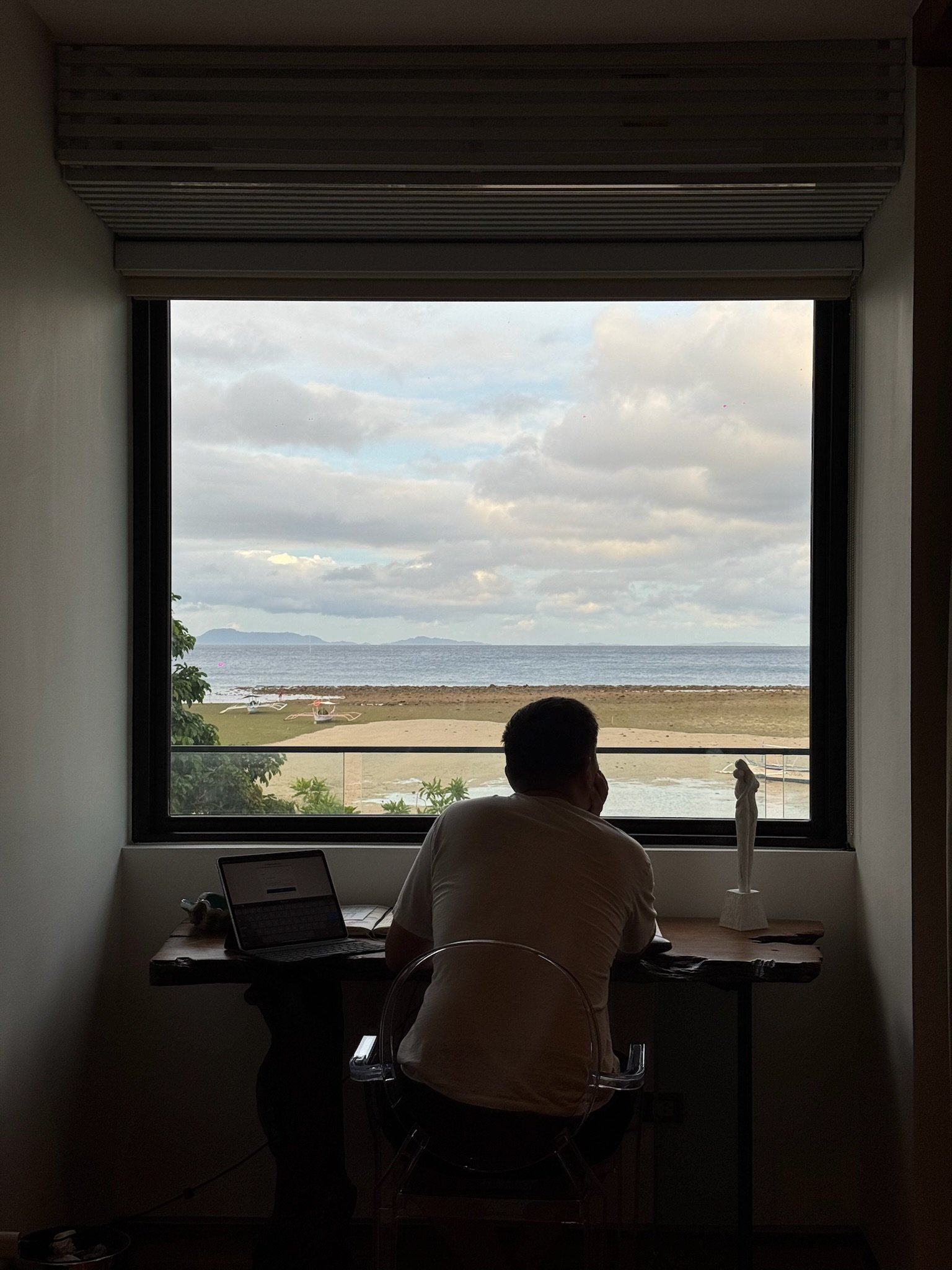 Person sitting at a desk with a laptop, looking out a large window at a beach and ocean view, with blue skies and distant islands.