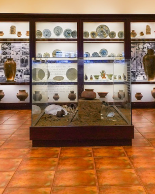 Museum display case with ancient pottery and artifacts on shelves and in a glass case, with tiled floor.