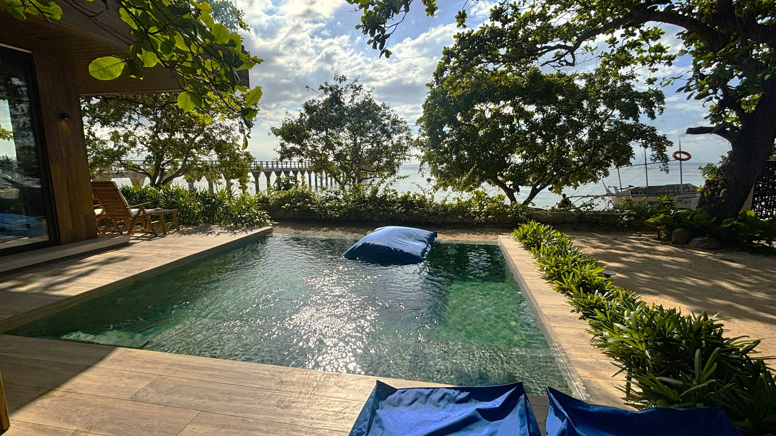 A small swimming pool with clear water and a deflated blue float near the center, surrounded by a wooden deck with lounge chairs. In the background, there are lush green trees, a view of the ocean, a boat with a life ring, and a partly cloudy sky.