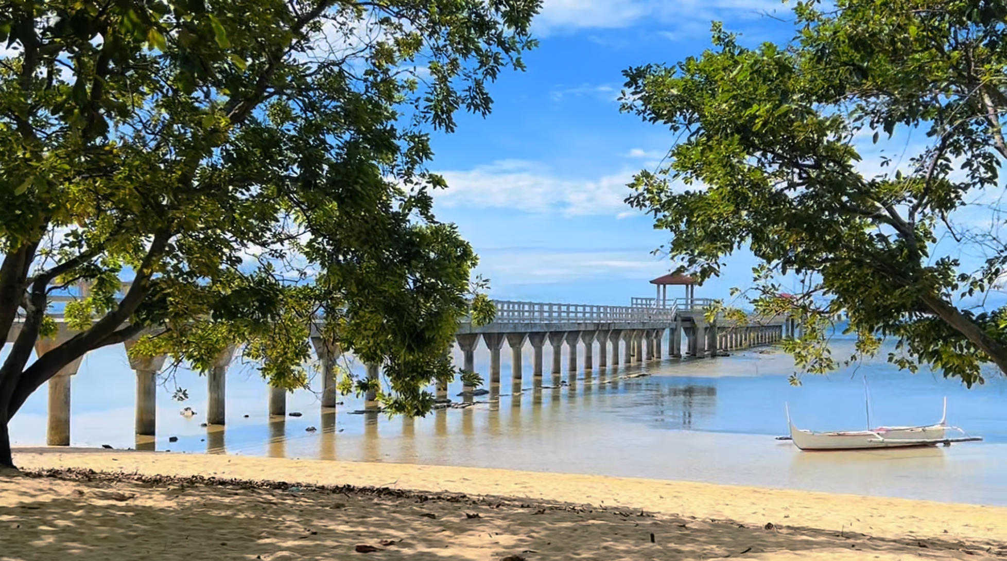 A tranquil beach scene with a sandy shore, green trees in the foreground, a long pier extending into calm blue water, and a small white boat floating near the shore. The sky is blue with some clouds.