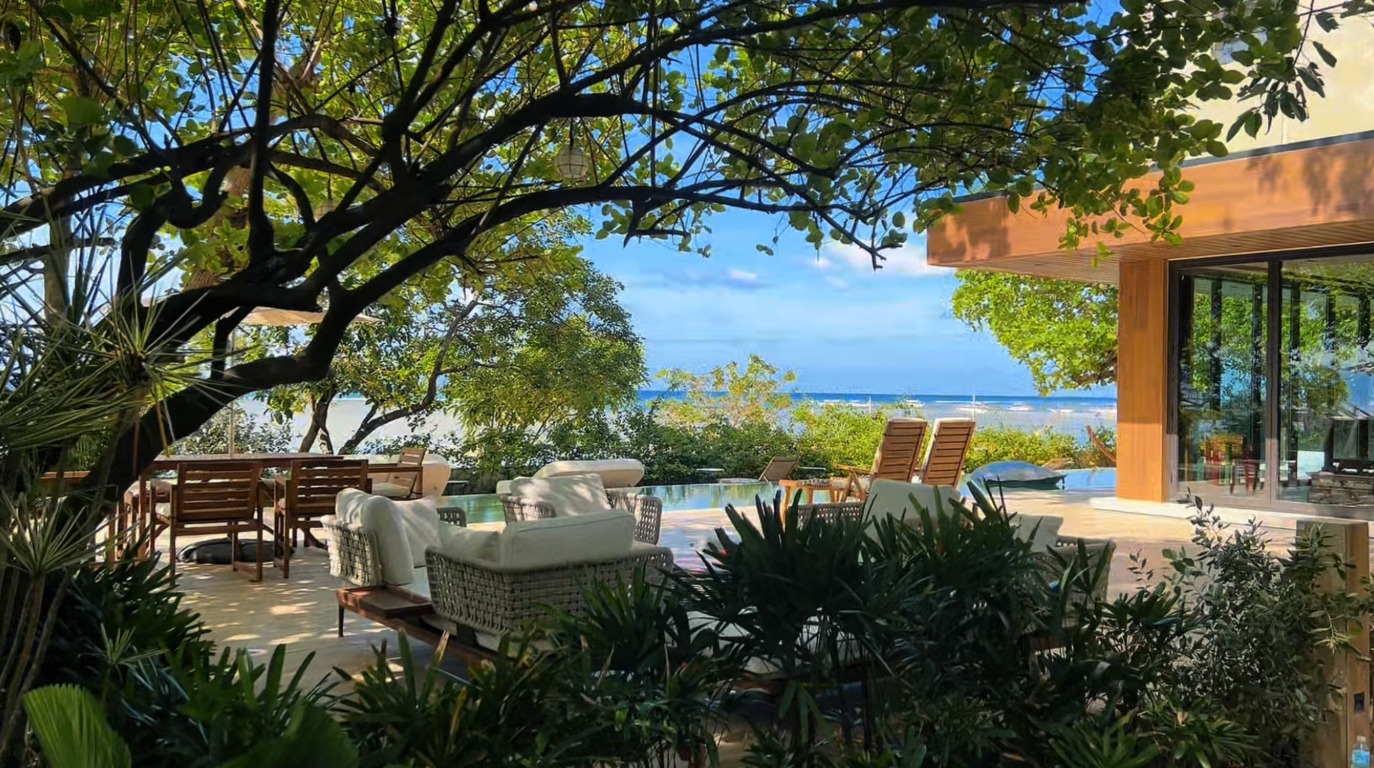 Outdoor patio area with lounge chairs, sofa, and dining table surrounded by trees and plants, overlooking a beach and ocean under a blue sky.